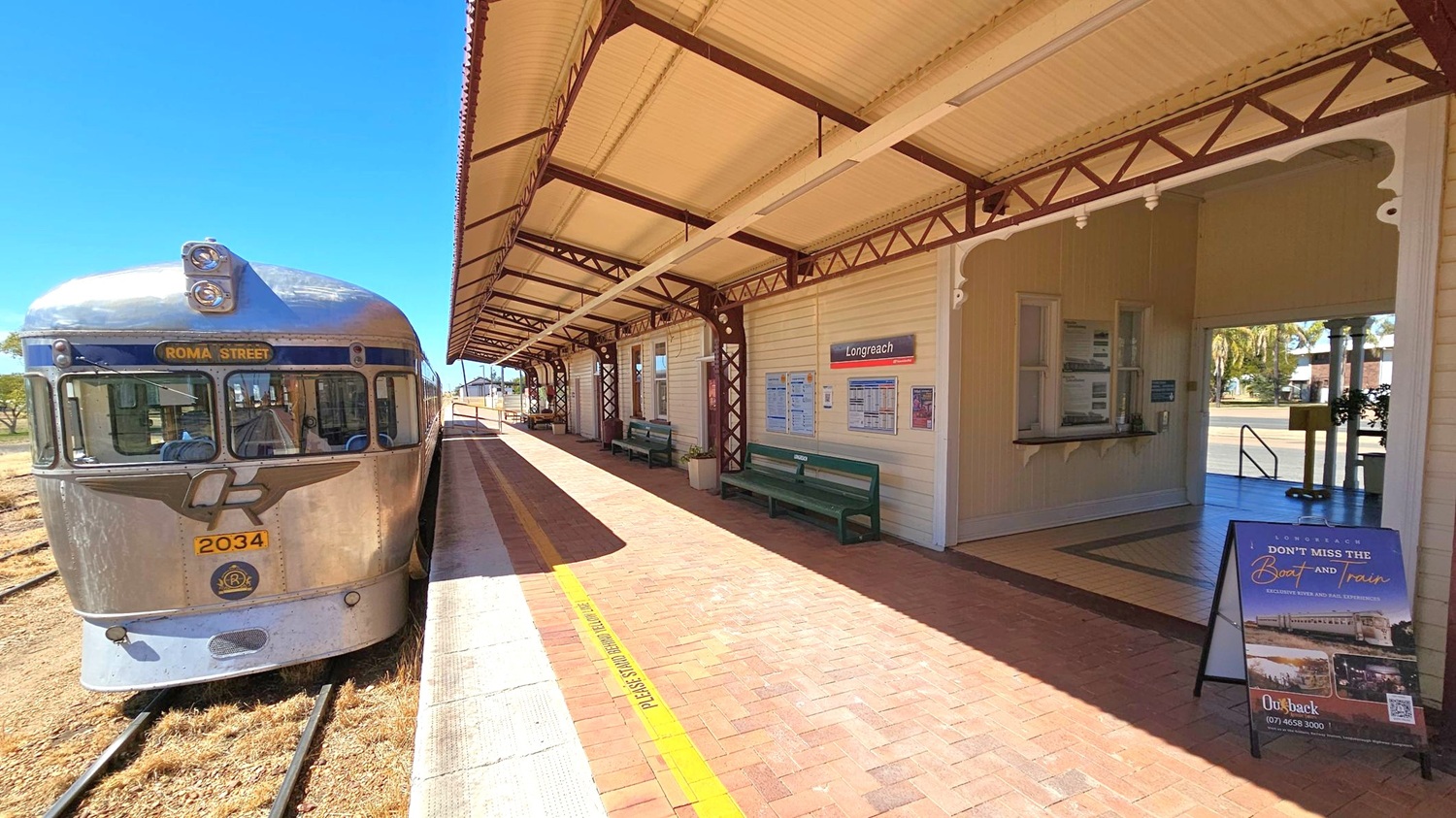 A silver train with the sign "Roma Street" is on the left, positioned on railway tracks. The platform is on the right, with a covered walkway supported by metal beams. The station building has a sign reading "Longreach" and features benches and information boards. A poster stand is near the entrance, which leads to an open area with visible trees in the background. The floor is paved with bricks, and the ceiling is made of corrugated metal.