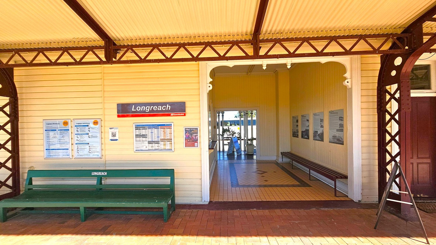 A railway station platform with a sign reading "Longreach" on a yellow wooden wall. Below the sign, there are several information boards and a green bench labelled "Longreach". To the right, an open passageway leads to an interior with tiled flooring and benches along the walls. The ceiling is corrugated, supported by decorative metal beams. The floor is brick, and there is a dark wooden door on the far right.