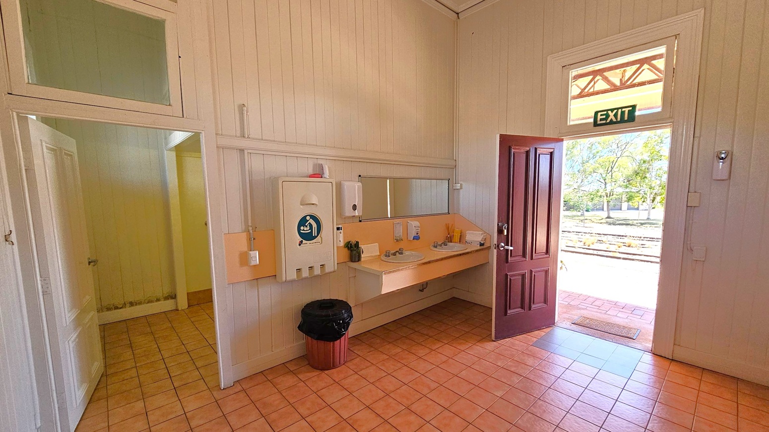 A restroom with light-coloured wooden walls and a tiled floor. On the left, there is a partially open door leading to another room. In the centre, a wall-mounted baby changing station, a mirror, and two sinks with soap dispensers are visible. On the right, a dark wooden door is open, leading outside. Above the door, an "EXIT" sign is displayed. Outside, trees and a paved area can be seen.