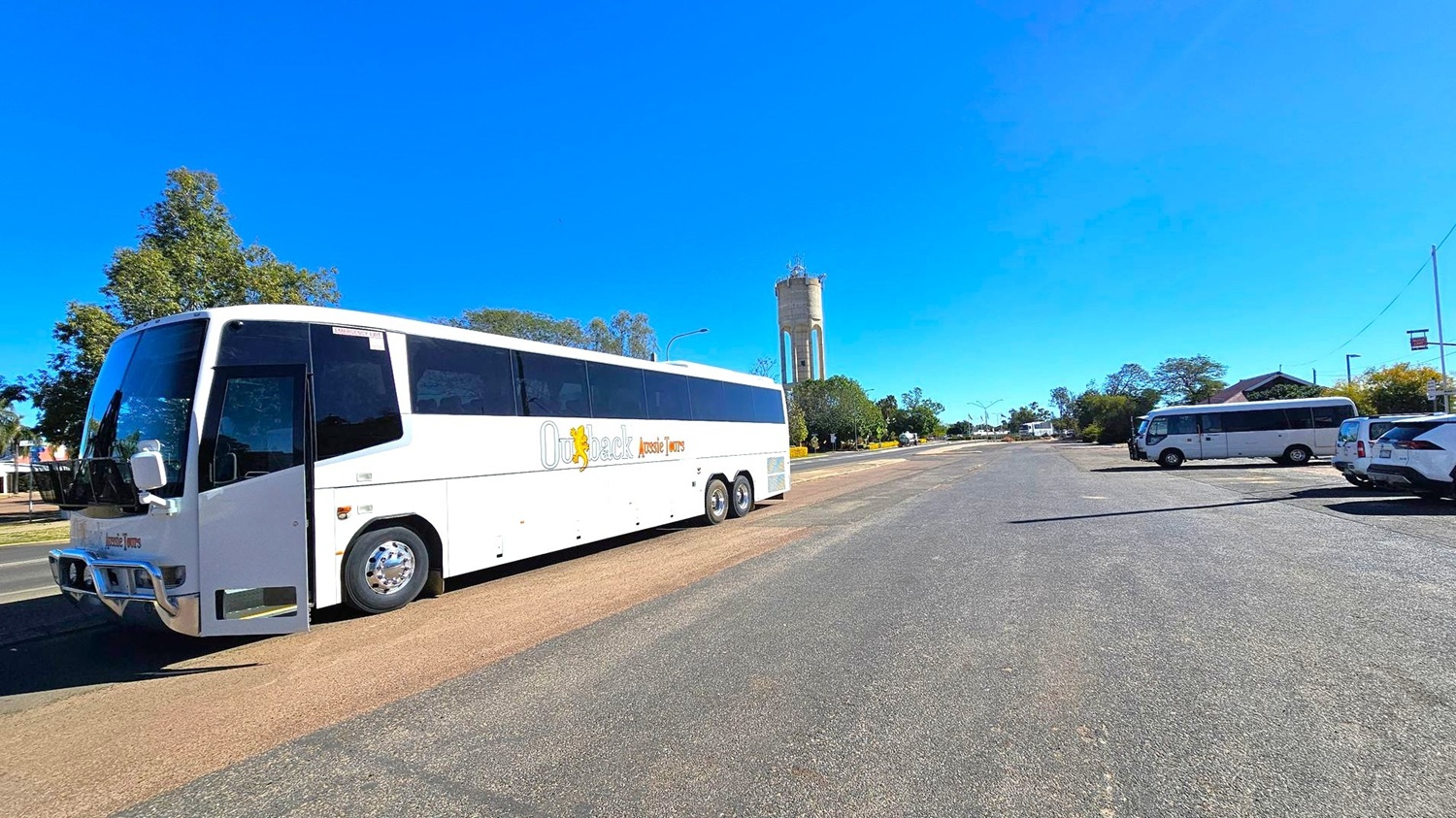 A large white tour bus is parked on the left side of a wide, open road. The bus has "Outback Aussie Tours" written on its side. In the background, a tall water tower is visible, surrounded by trees. On the right side, a smaller bus and a car are parked along the road. The area is spacious with clear skies above.