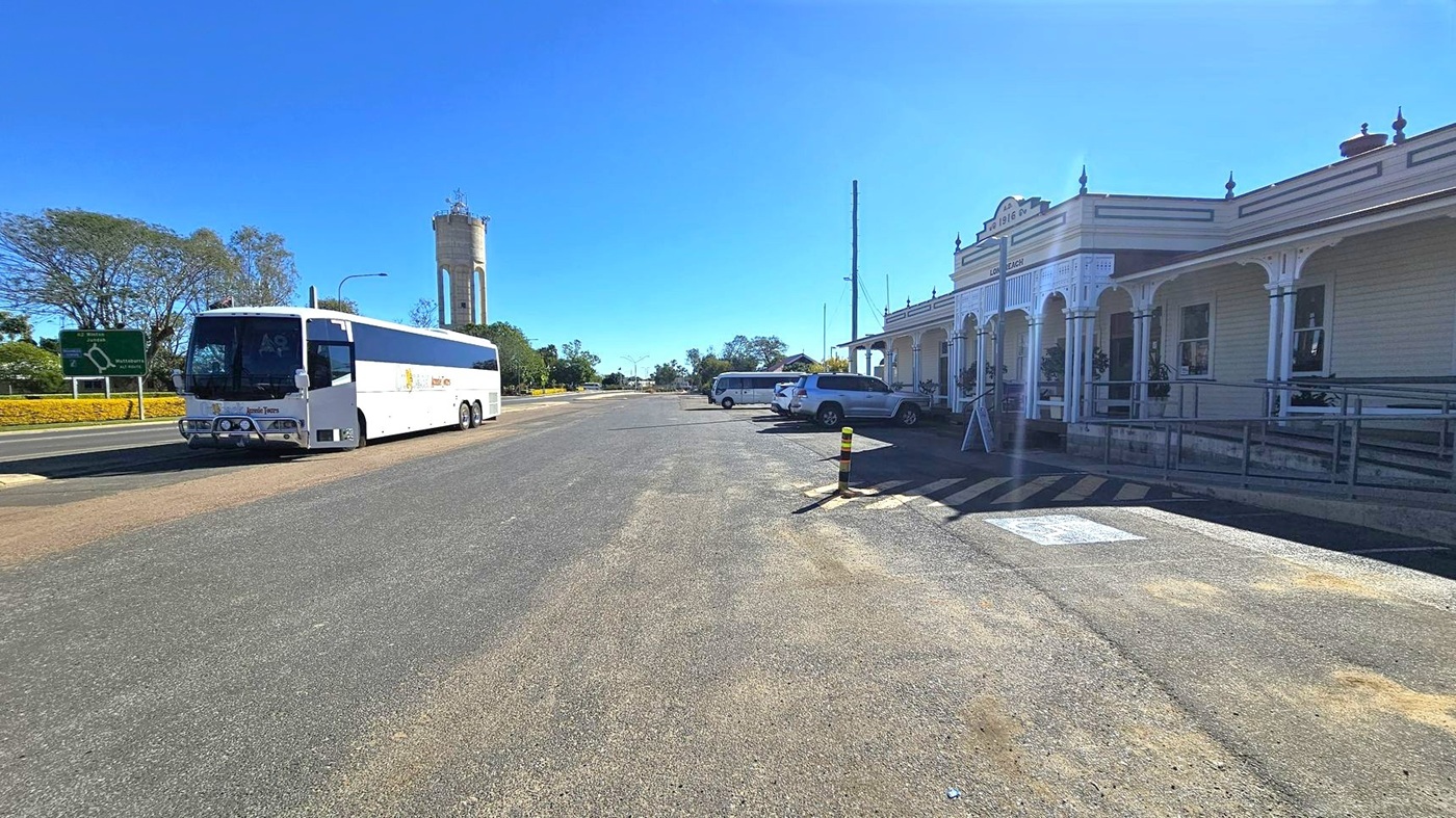 A wide street with a white bus parked on the left side. In the background, a tall water tower is visible. On the right, there is a historic building with a veranda and decorative columns, featuring a sign with the year 1916. Several cars are parked in front of the building. The pavement has a marked disabled parking space, and there are some bollards near the building. Trees and a road sign are visible in the distance.