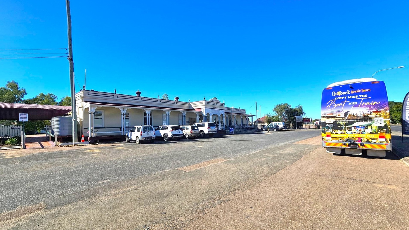 A street scene with a row of historic buildings on the left, featuring a covered walkway and several parked white vehicles. A tall utility pole stands nearby. On the right, a large tour bus with colourful graphics and text advertising "Outback Aussie Tours" is parked. The road is wide with patches of grass and dirt. Trees and additional vehicles are visible in the background. The sky is clear and blue.