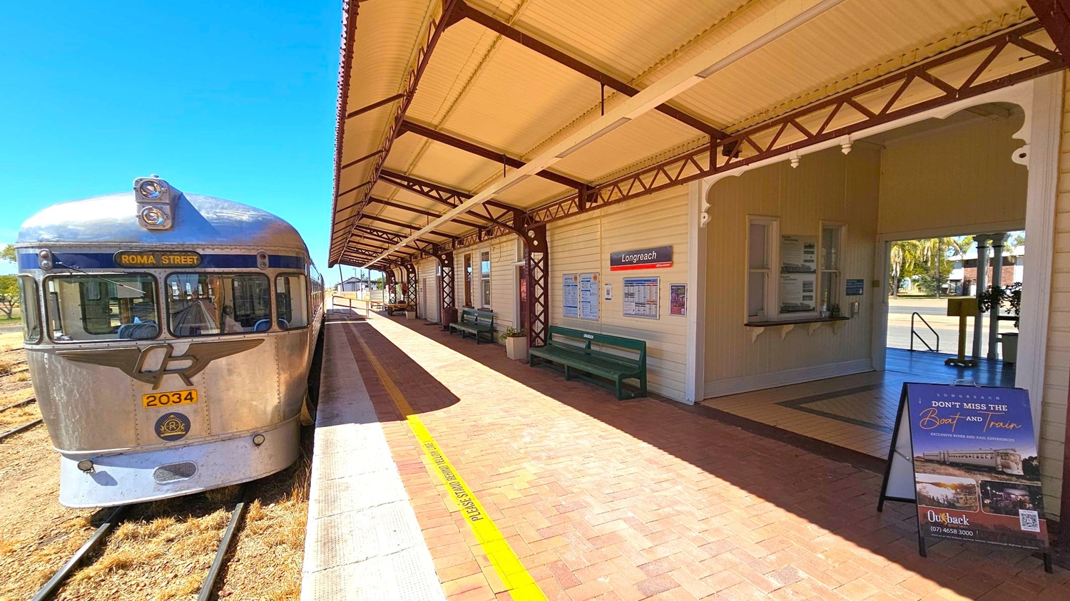 A vintage silver train with "Roma Street" displayed on the front is on the left, positioned on railway tracks. The platform is on the right, with a covered walkway featuring a corrugated roof and decorative metal supports. Benches and a sign reading "Longreach" are visible along the wall. A poster stand is near the entrance to a building, which has a ticket window and leads to an outdoor area with trees. The floor is paved with red bricks.