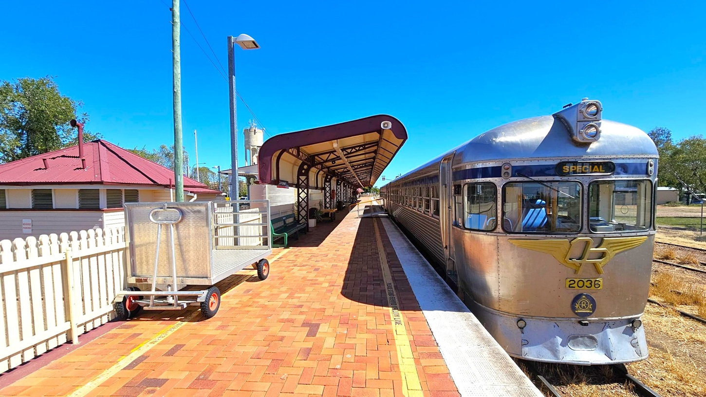 A vintage silver train labelled "Special" is on the right, positioned on a railway track beside a platform. The platform is paved with reddish-brown bricks and features a covered waiting area with a maroon and cream roof. On the left, there's a white picket fence and a metal trolley with red wheels. A building with a red roof is visible behind the fence. In the background, a water tower and trees can be seen.