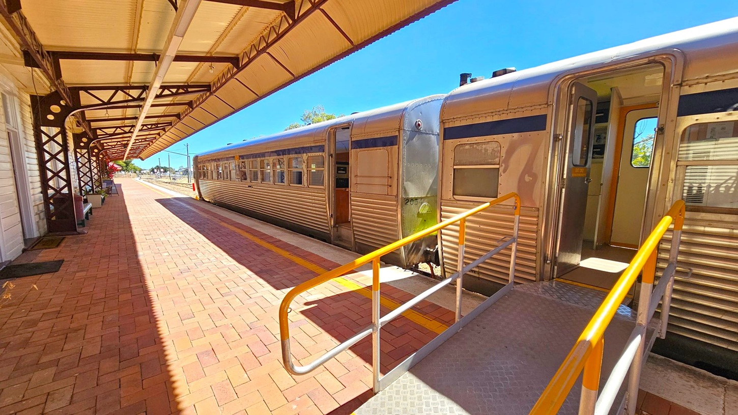A train station platform with a metal train on the right. The train has an open door with a ramp leading to it, featuring yellow handrails. The platform is paved with red bricks and has a covered roof supported by metal beams on the left. Benches are placed along the wall of the station building. The sky is clear and blue, and there are trees visible in the background.