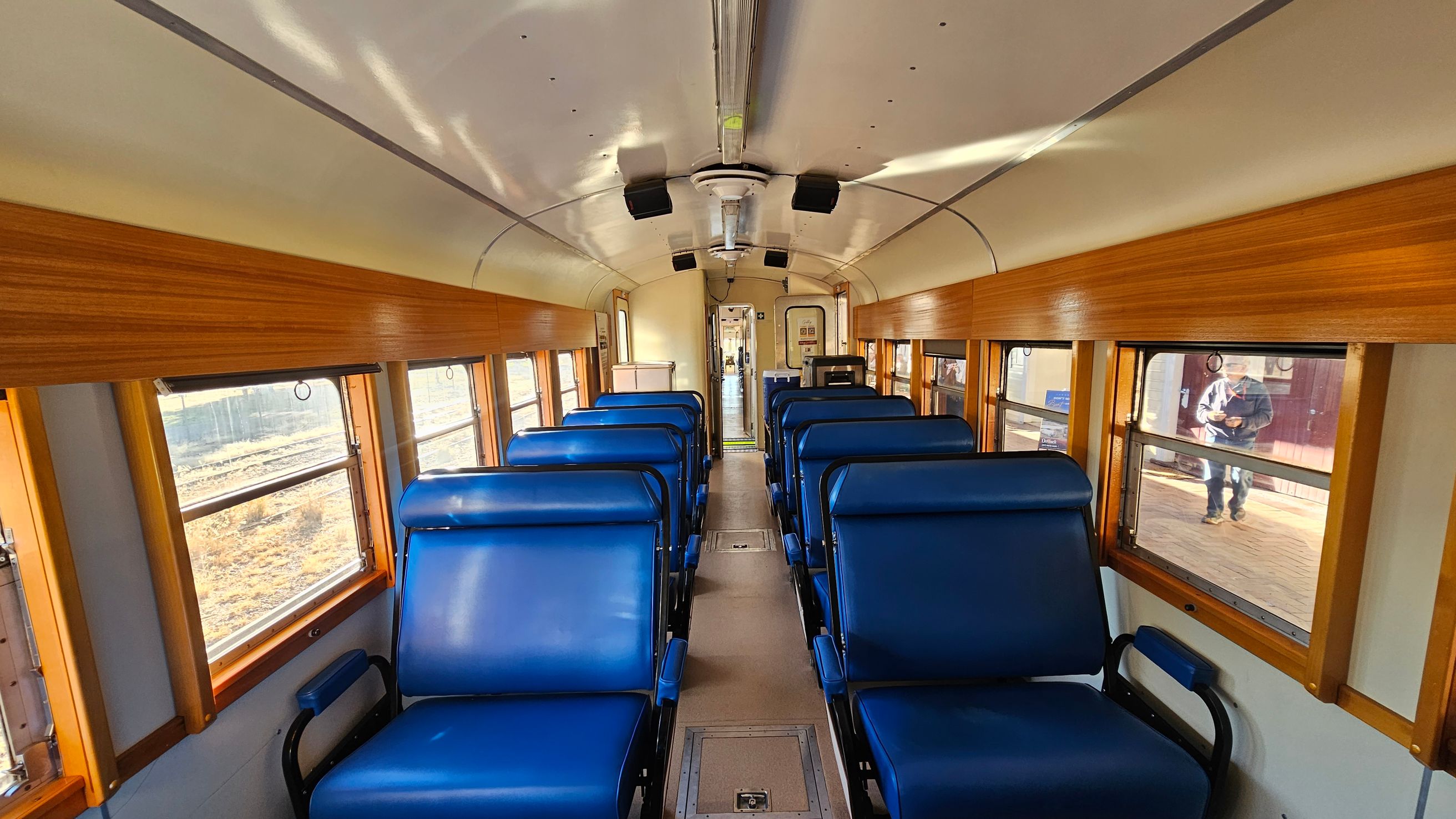 Inside a vintage train carriage with blue cushioned seats arranged in pairs on either side of a central aisle. The walls are lined with wooden panels, and large windows allow natural light to enter. The ceiling is curved and fitted with lights and speakers. At the far end, a door leads to the next carriage. A person is visible outside through the window on the right. The floor is a light-coloured material, and the overall atmosphere is bright and spacious.