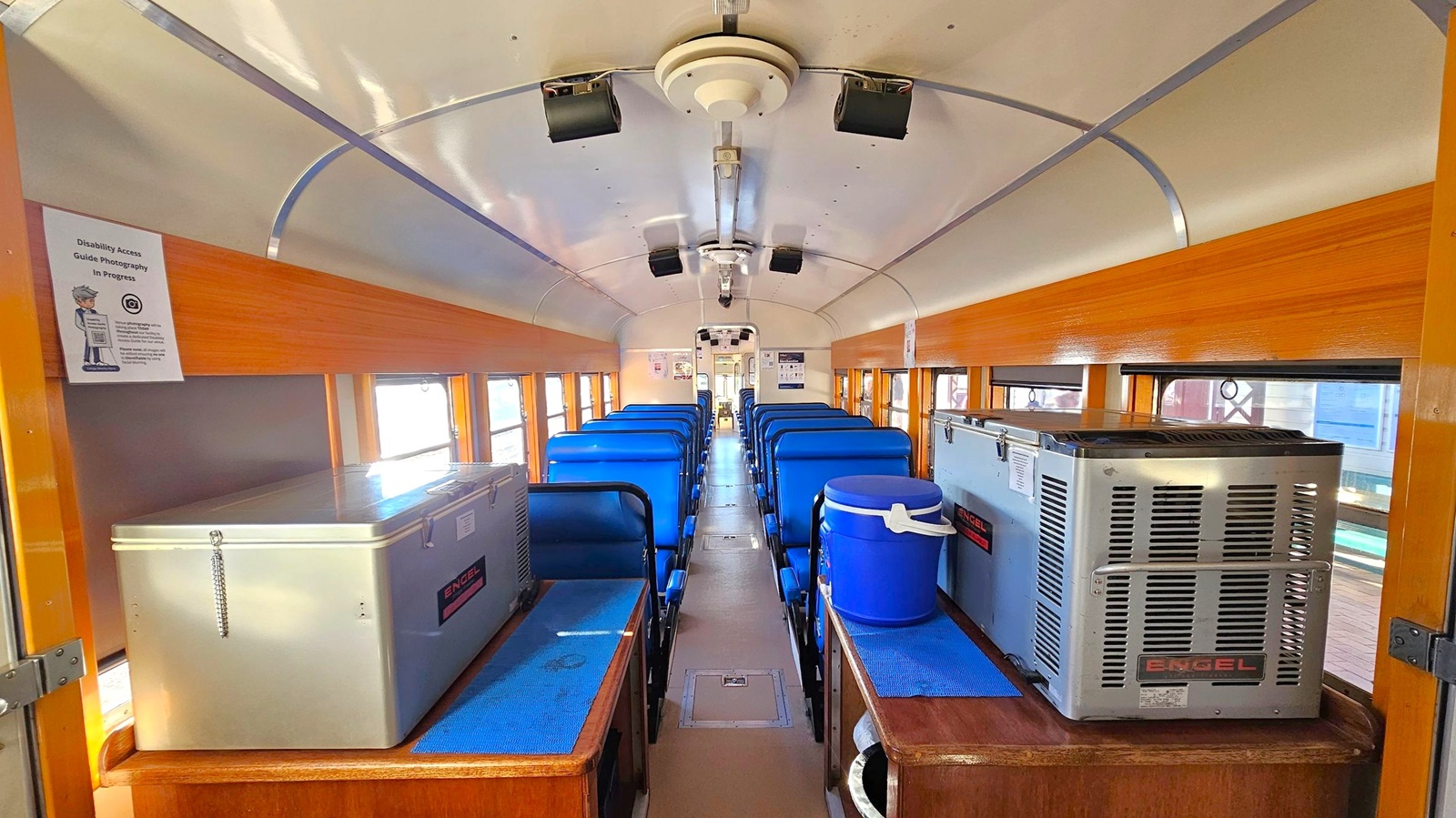 Inside a train carriage with a white ceiling and wooden panelling along the walls. Blue cushioned seats are arranged in rows on either side of a central aisle. On the left, a large white cooler is placed on a wooden surface. On the right, a silver cooler and a blue water dispenser are positioned similarly. Windows line both sides, allowing natural light to illuminate the interior. A sign on the left wall mentions disability access.