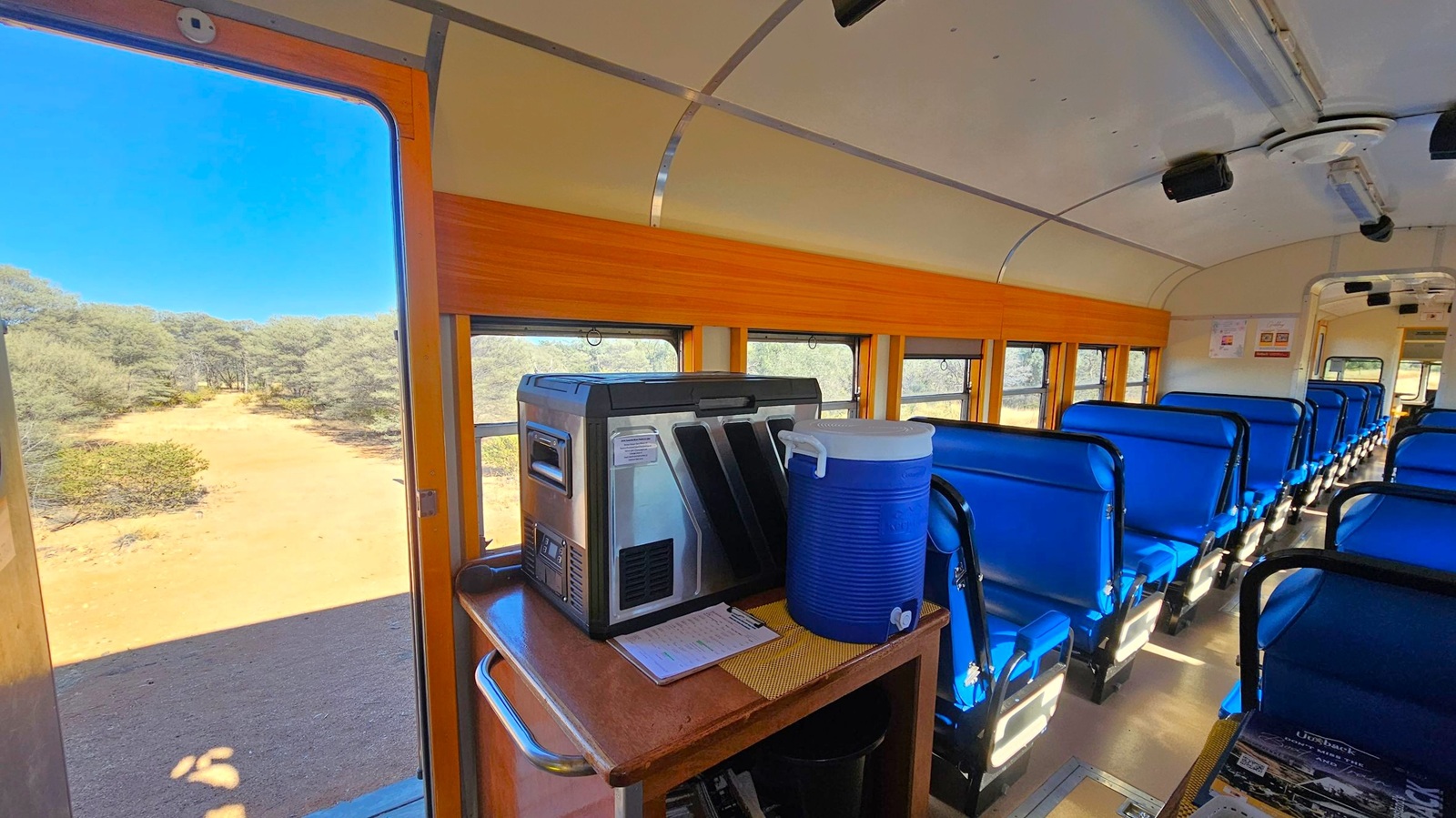 Inside a train carriage with blue seats arranged in rows on the right. A wooden panel runs along the windows. On the left, an open door reveals a view of a sandy path and trees outside. In the foreground, a table holds a large blue cooler and a metallic appliance. The ceiling is curved with lighting fixtures, and the floor is light-coloured. Posters are visible on the far wall.