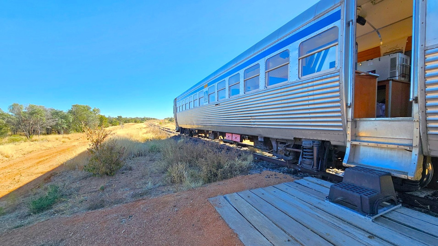 A silver train carriage with blue accents is on the right, with an open door revealing an interior with a desk and air conditioning unit. A small black step is placed on a wooden platform leading to the door. On the left, a dirt path runs alongside the train, bordered by dry grass and scattered bushes. In the background, there are trees and a clear horizon. The ground is a mix of gravel and dirt.