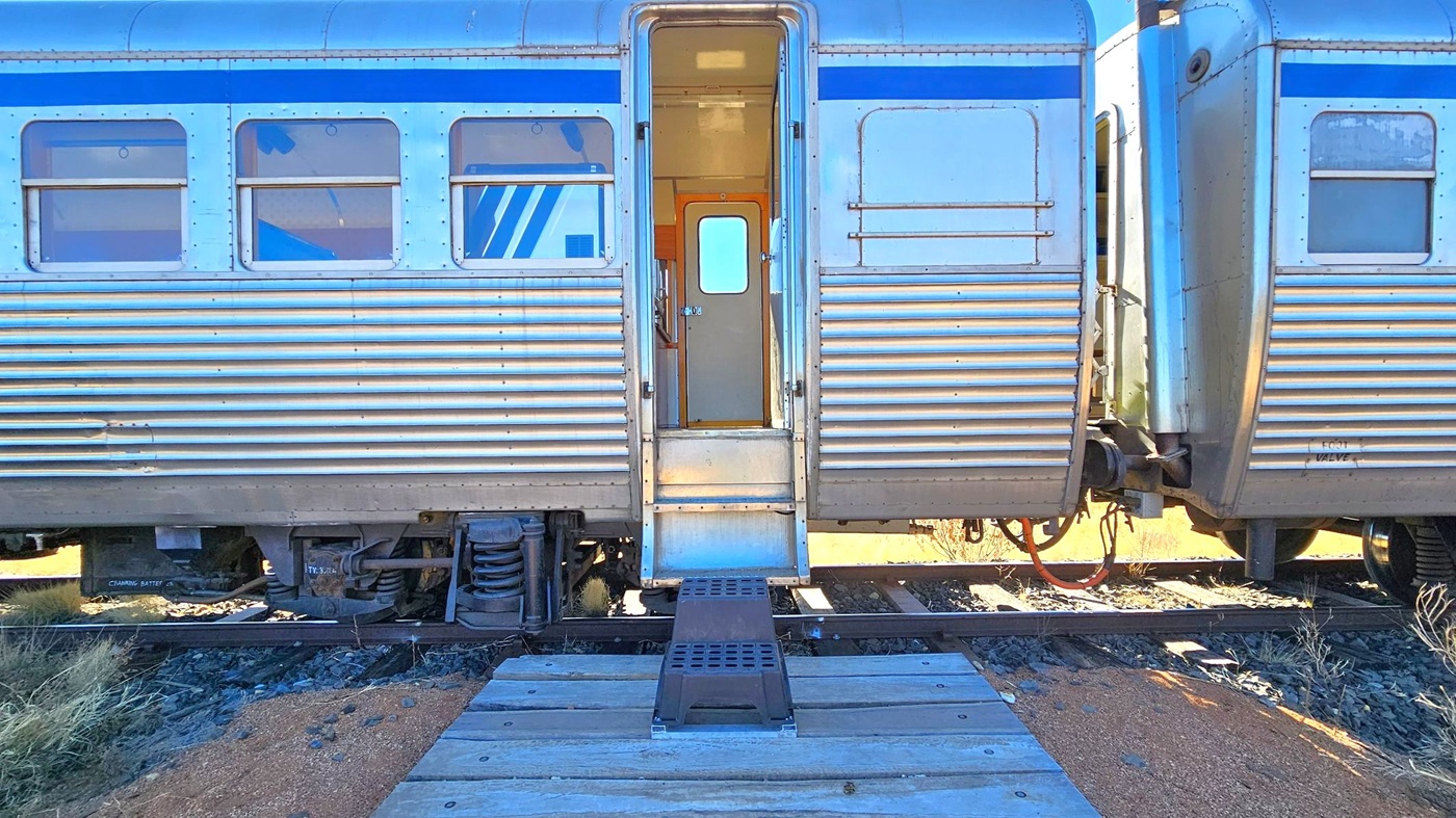A silver train carriage with horizontal ridges and a blue stripe near the top. The central door is open, revealing a narrow interior passageway. A small set of steps leads down to a metal platform, which connects to a wooden walkway on the ground. The train is on tracks surrounded by gravel and sparse grass. Another carriage is partially visible on the right, connected to the first.
