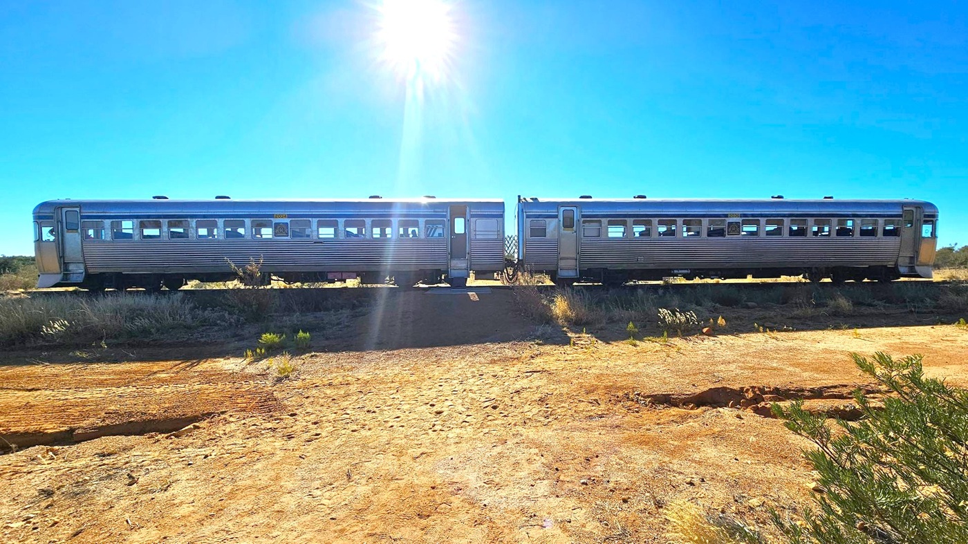 A silver train with two carriages is on a track elevated above a dry, sandy terrain. The train has a sleek, metallic exterior with several windows. The sun is positioned high in the sky, casting a bright light and creating a lens flare effect. Sparse vegetation is visible in the foreground, with some small bushes and patches of grass. The background is a clear blue sky.