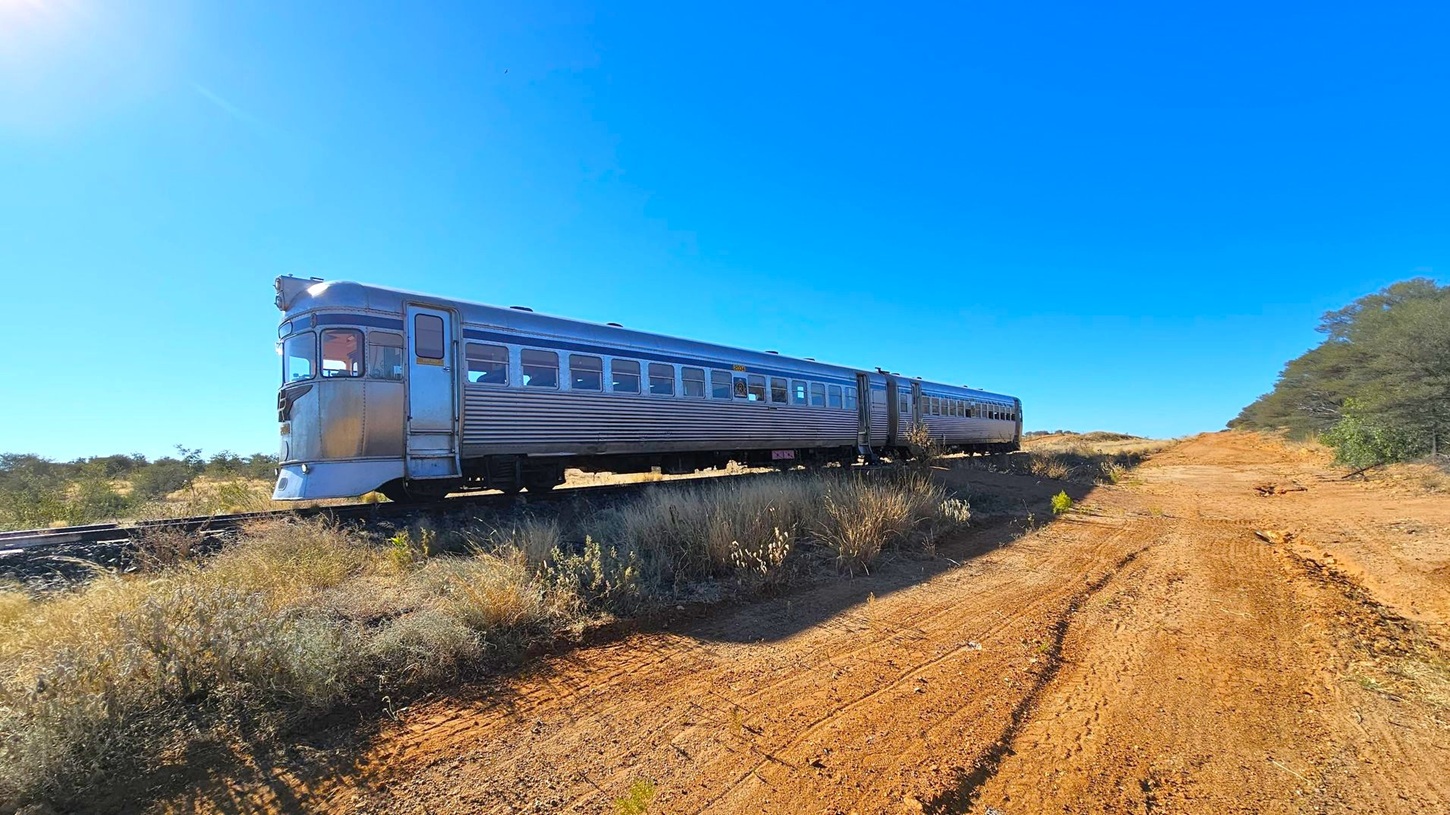 A silver train with multiple carriages is on the left, travelling along a railway track. The train has a vintage design with large windows. On the right, there is a dirt path with visible tyre tracks, leading into the distance. The foreground is covered with dry grass and shrubs, while the background features a clear blue sky and sparse trees on the right.