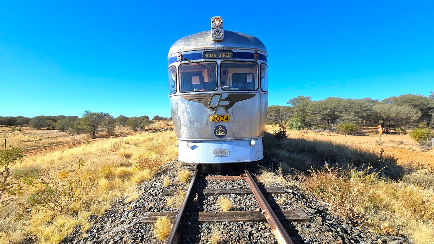 A silver train with the number 2034 is positioned on railway tracks, facing forward. The train has a rounded front with large windows and a sign reading "ROMA STREET." The surrounding landscape is dry, with sparse grass and bushes on either side of the tracks. In the background, there are scattered trees and a person in an orange outfit standing to the right. The sky is clear and blue.