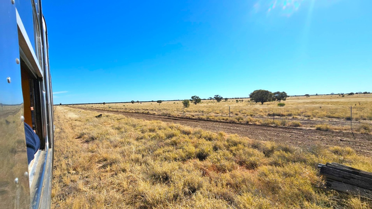 A view from a train window on the left, showing a vast, open landscape. The foreground features dry grass and a dirt path running parallel to the train. In the background, scattered trees dot the expansive, flat terrain under a clear blue sky. A fence runs alongside the path, and a few wooden planks are visible in the lower right corner.