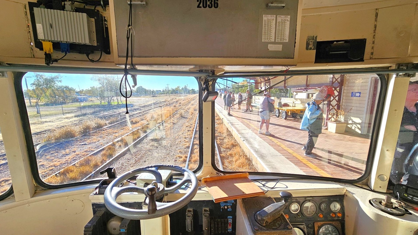 View from inside a train cab looking out through the front windows. The left side shows railway tracks and dry grass. The right side features a platform with people walking, some wearing hats. The cab interior includes a steering wheel, control panel with dials, and a clipboard. Overhead, there are wires and equipment. The ceiling is beige, and the floor is not visible. Trees and a fence are in the background.
