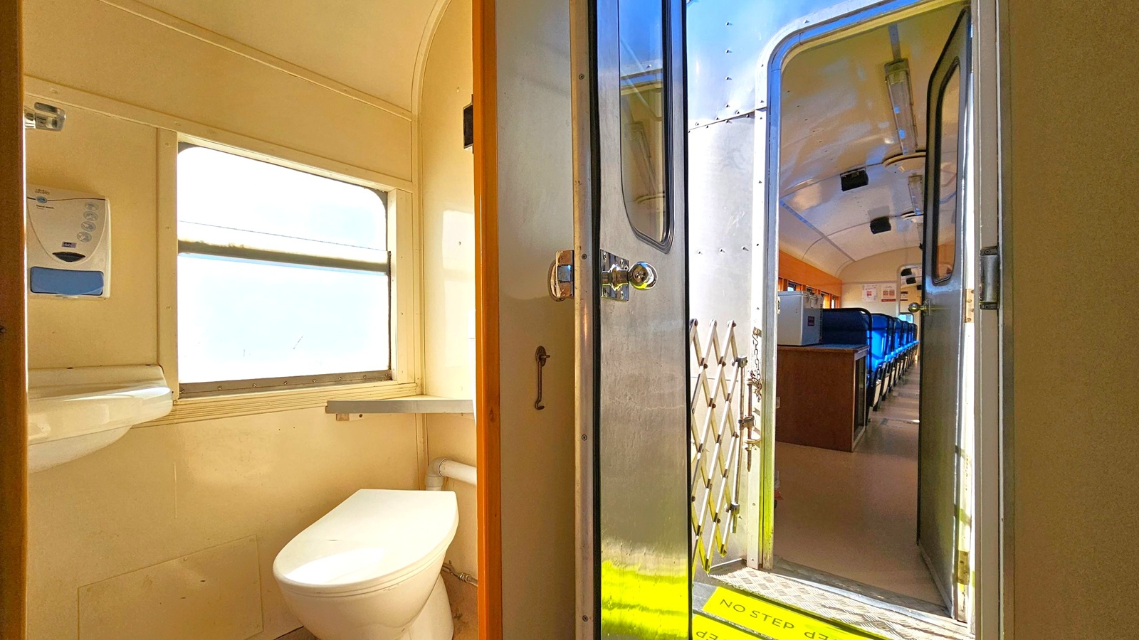 On the left, there's a small train bathroom with a white toilet and a window above it. A soap dispenser is mounted on the wall. In the middle, a metal door with a handle is partially open. On the right, the interior of a train carriage is visible, featuring rows of blue seats. The floor is light-coloured, and the ceiling has fluorescent lights. A yellow "No Step" sign is on the floor near the door.