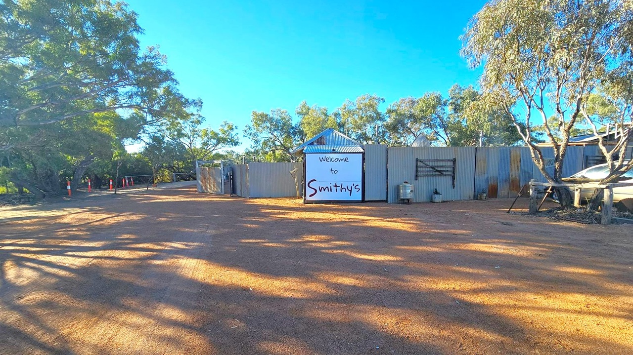 A dirt path leads to a corrugated metal fence with a large sign reading "Welcome to Smithy's" in the centre. Trees with sparse foliage are on both sides, casting shadows on the ground. To the right, a vehicle is partially visible behind the fence. The sky is clear and blue.