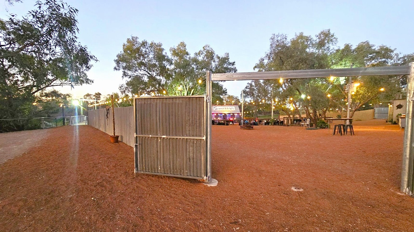 A rustic outdoor area with a dirt ground and a corrugated metal gate in the centre. On the right, there are string lights hanging above, illuminating tables and stools scattered around. In the background, a sign reads "Smithy's" above a structure with people gathered nearby. Trees surround the area, and a wooden fence runs along the left side, with more trees visible beyond it.