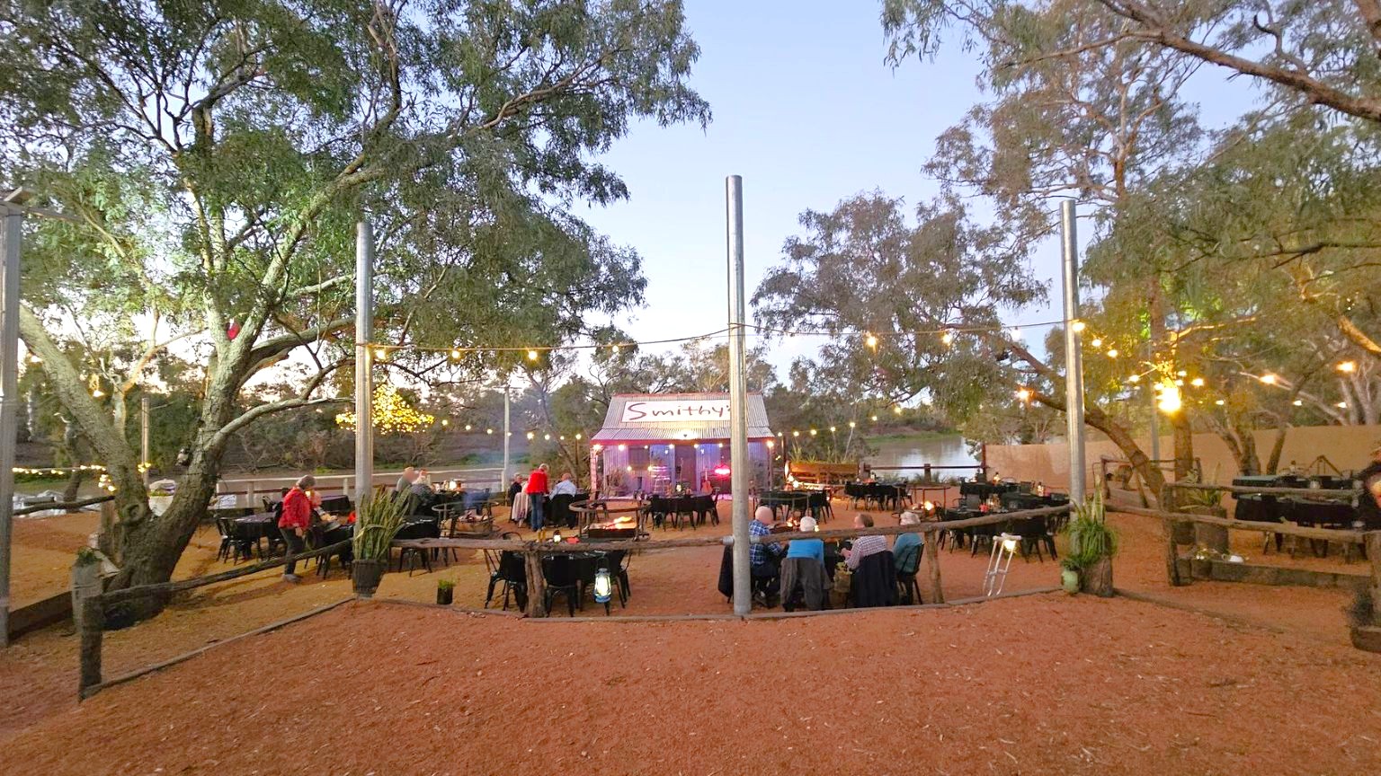 Outdoor dining area with wooden tables and chairs on a dirt ground. In the centre, a small building with a corrugated metal roof displays the name "Smithy's". String lights are hung between tall poles, illuminating the area. Large trees surround the space, and a river is visible in the background. People are seated at tables, and some are standing near the building. The atmosphere is relaxed and rustic.
