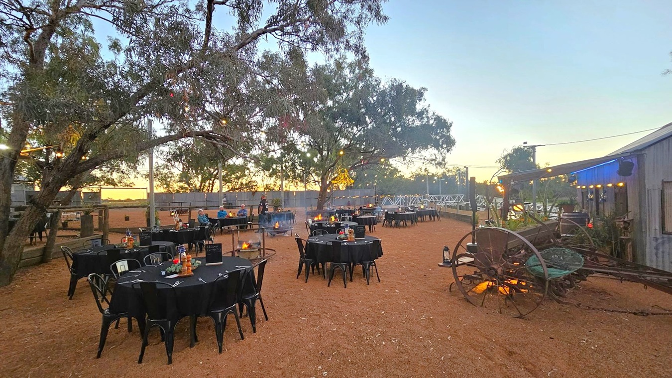 Outdoor dining area with round tables covered in black tablecloths, set with cutlery and lanterns. On the left, large trees with string lights create a canopy. In the centre, a few people are seated at a table. On the right, a rustic wooden building with a blue-lit roof and an old wagon. The ground is covered in reddish-brown dirt, and there are fire pits near some tables. A wooden fence runs along the background, with a sunset visible through the trees.