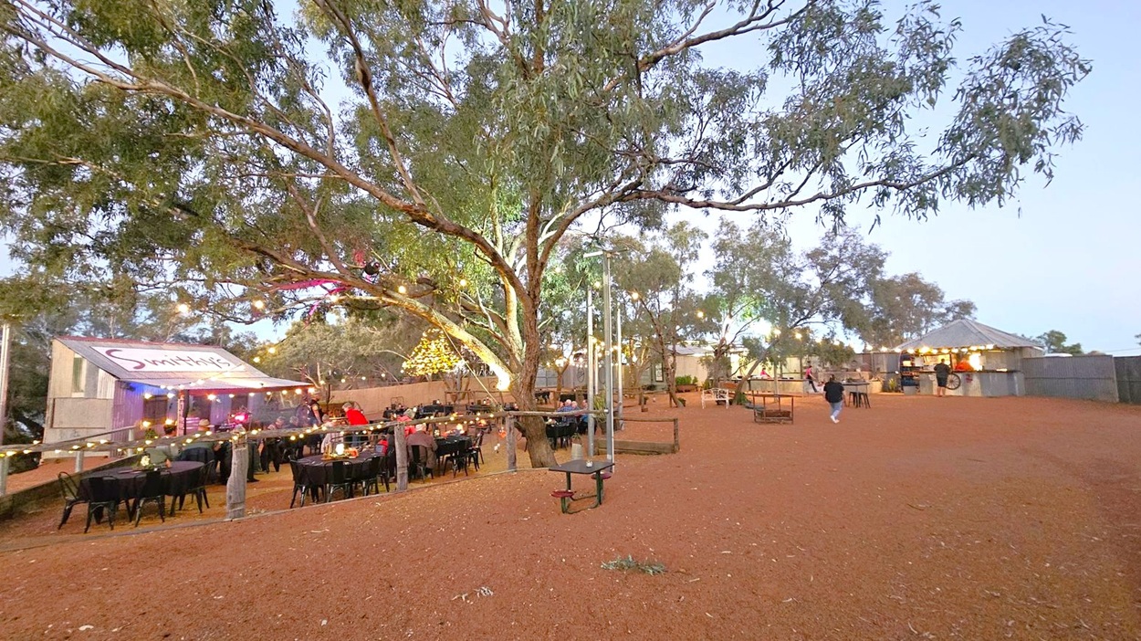An outdoor dining area with a rustic setting. On the left, there's a building with a sign that reads "Smith's" and tables with black tablecloths and chairs beneath a large tree adorned with string lights. In the centre, more tables are scattered under trees, also decorated with lights. On the right, a small structure with a conical roof is visible, possibly a bar or food stand. The ground is covered with reddish-brown gravel, and the area is enclosed by a wooden fence.