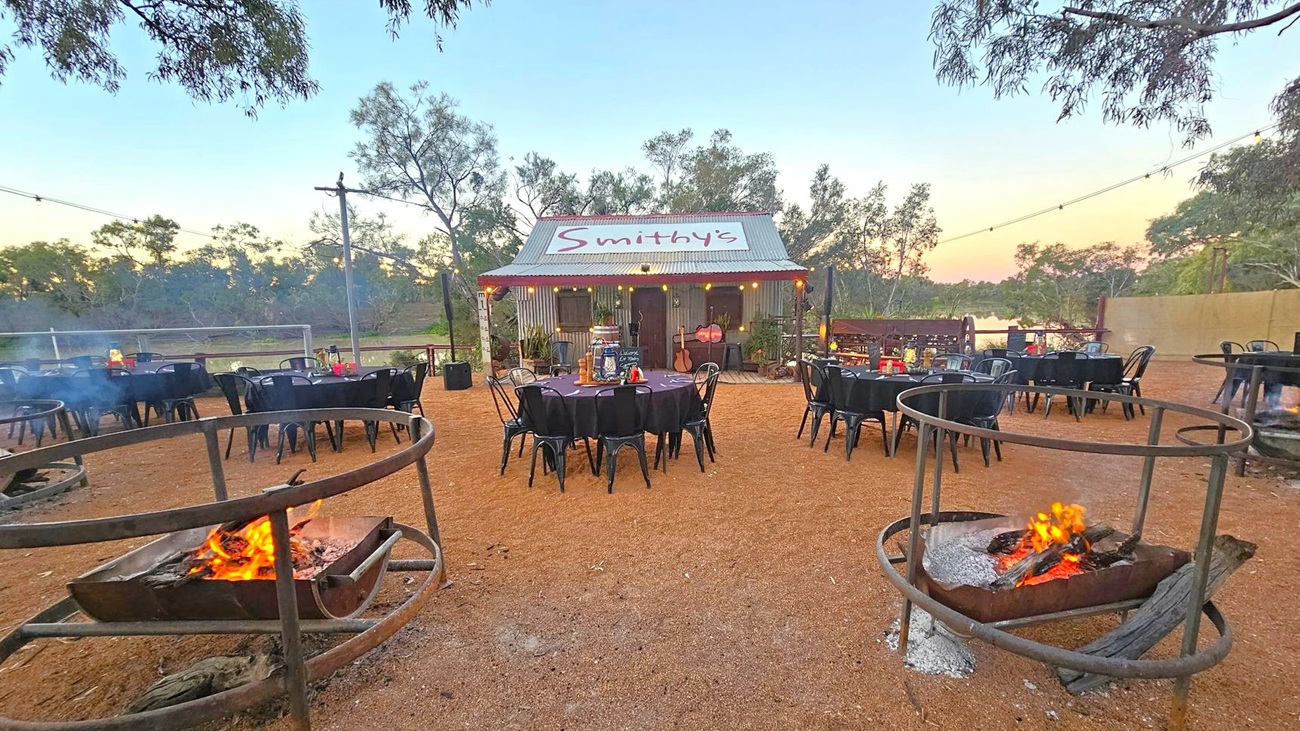 A rustic outdoor setting with a small corrugated metal building labelled "Smithy's" in the centre. In the foreground, there are two circular fire pits with burning logs. Several round tables with black tablecloths and chairs are arranged around the area. String lights are hung above, and trees are visible in the background. The ground is covered with reddish-brown gravel.