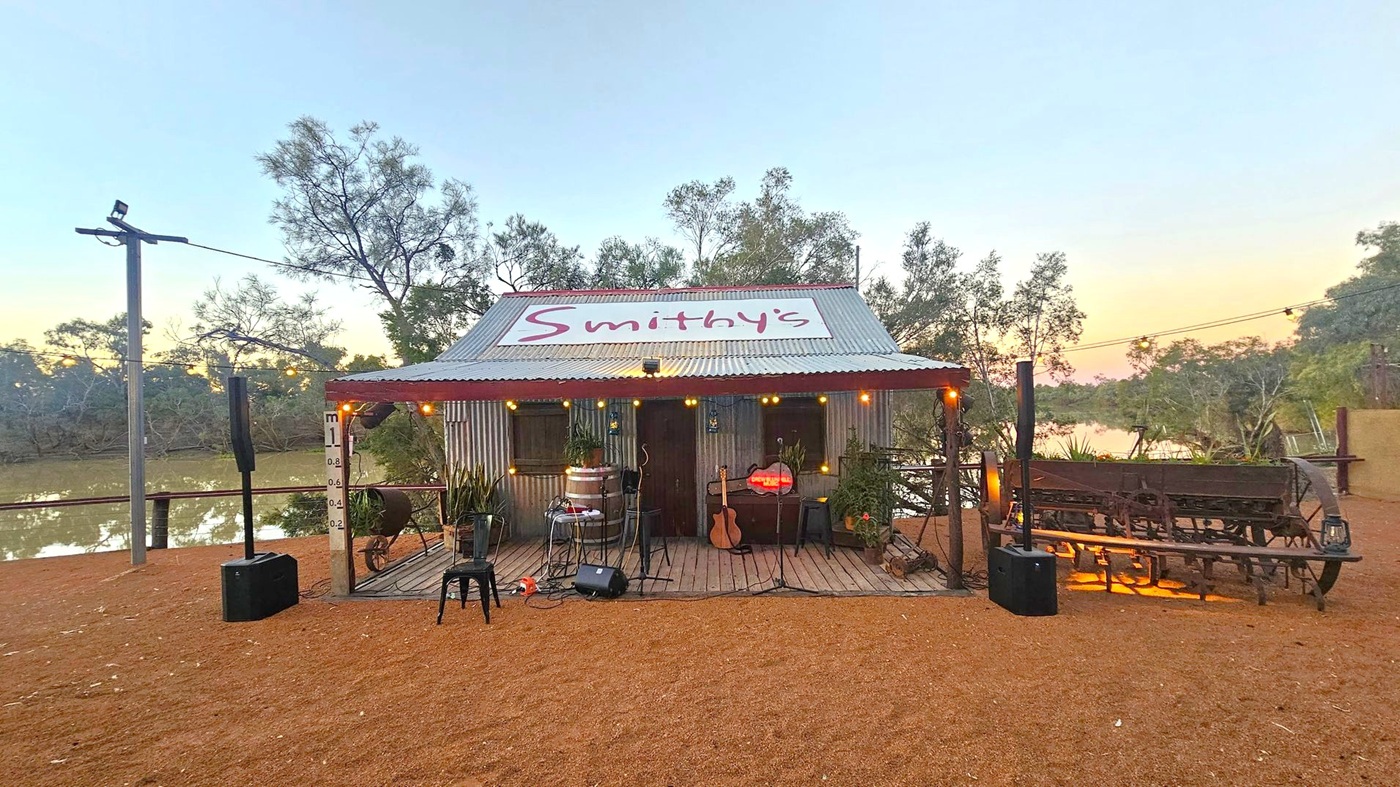 A rustic structure with a corrugated metal roof and a sign reading "Smithy's" on top. The building is adorned with string lights and has a small stage area with musical equipment, including a guitar and microphone stands. In front, there are a few chairs and a barrel. The ground is covered with reddish-brown gravel. To the right, there's an old wooden cart. Trees and a body of water are visible in the background, with a soft sunset glow.