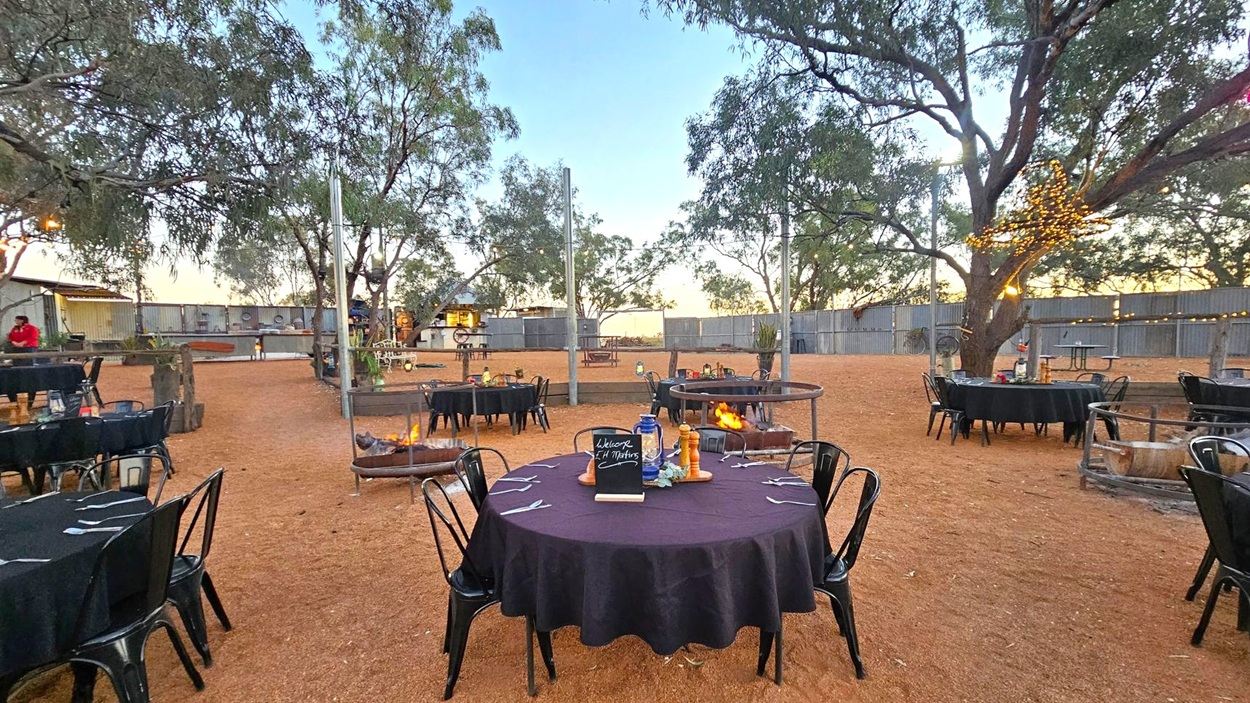 Outdoor dining area with round tables covered in black tablecloths, surrounded by black chairs. In the centre, a table displays a small chalkboard and condiments. Fire pits are scattered around, with one in the foreground. Trees with string lights are visible, and a wooden fence encloses the space. The ground is covered in reddish-brown dirt, and there are additional tables and a structure in the background.