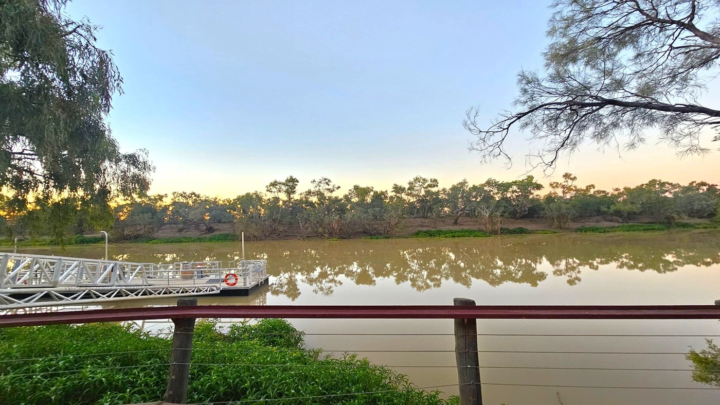 A calm river with a reflection of trees along the opposite bank. In the foreground, a metal railing and a small dock with a lifebuoy are visible. The dock extends into the water from the left. Trees with sparse branches frame the scene on both sides. The water is a muddy brown, and the sky has a gradient of light blue to pale orange near the horizon. Green foliage is visible near the railing.