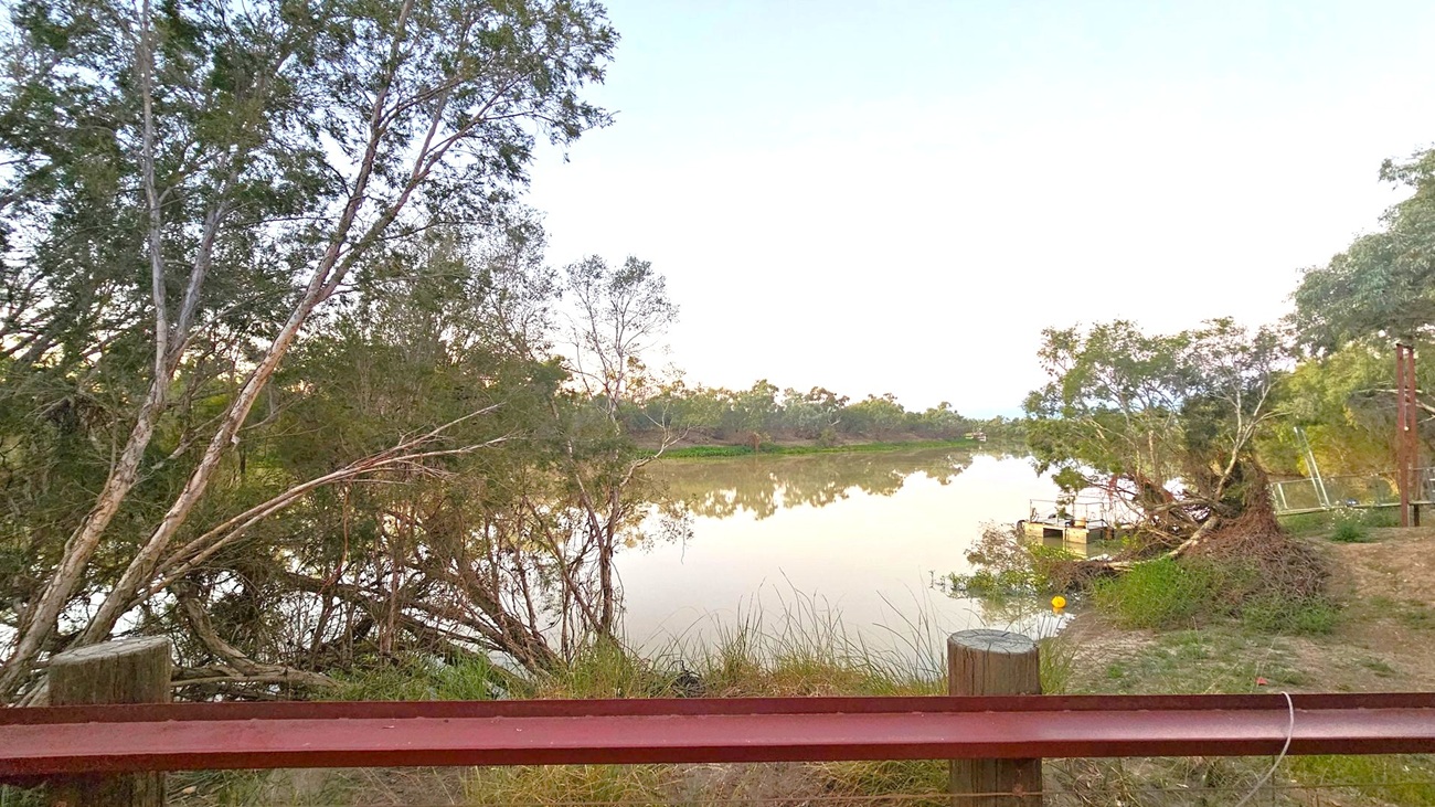 A calm river flows through the centre, reflecting the surrounding trees. On the left, tall trees with dense foliage lean over the water. On the right, a small boat is moored near the riverbank, surrounded by more trees and shrubs. A rusty metal railing runs across the foreground, with wooden posts supporting it. The background features a line of trees along the opposite bank, under a clear sky.