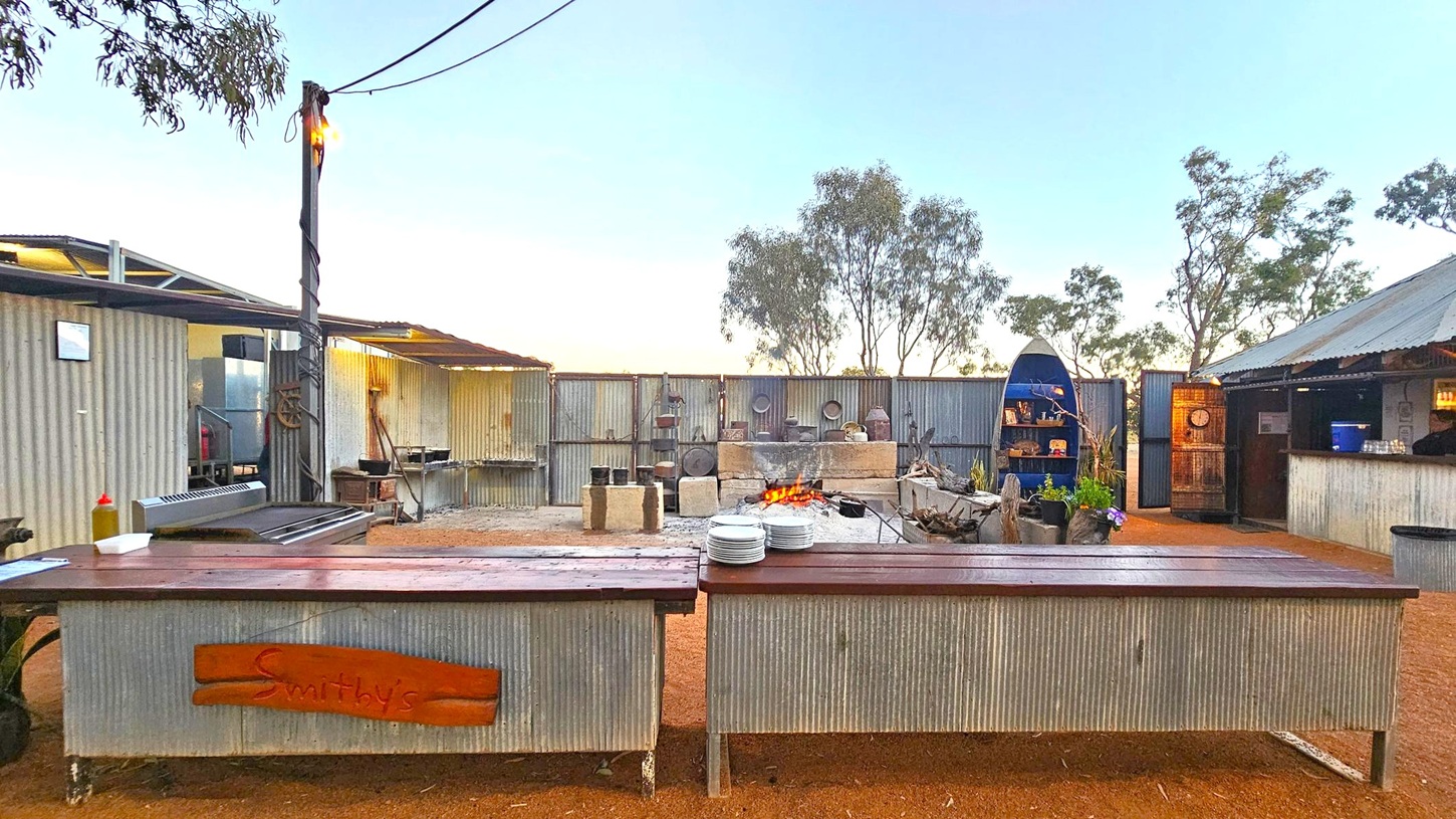 An outdoor kitchen area with corrugated metal structures. In the foreground, two long tables with a stack of plates and a bottle of sauce. The middle features a fire pit surrounded by stone blocks, with cooking pots nearby. In the background, a fence with various items hanging and a blue boat-shaped shelf with decorations. Trees are visible beyond the fence. The ground is dirt, and the area is lit by a single overhead bulb.