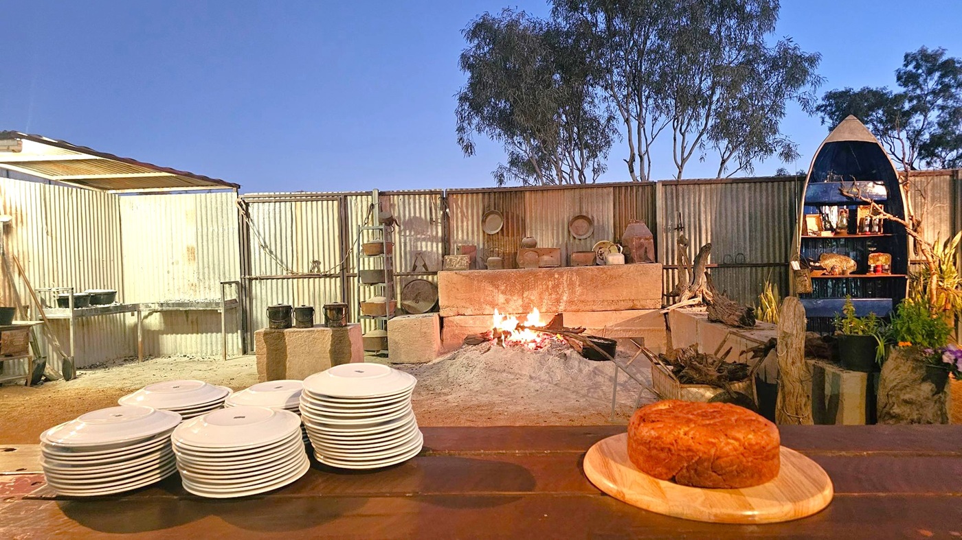 A rustic outdoor setting with a central fire pit surrounded by corrugated metal walls. On the left, stacks of white plates are placed on a wooden surface. In the foreground, a round loaf of bread sits on a wooden board. To the right, a boat-shaped shelf holds various items, and there are plants and logs nearby. The background features trees silhouetted against the evening sky.