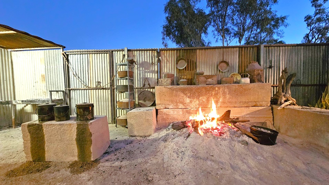 A rustic outdoor setting with a corrugated metal wall in the background. In the centre, a fire burns on a sandy surface, surrounded by large stone blocks. To the left, three black pots sit on a stone block. On the right, various metal containers and kettles are arranged on shelves and ledges. A tree is visible above the wall, and the ground is covered in sand.
