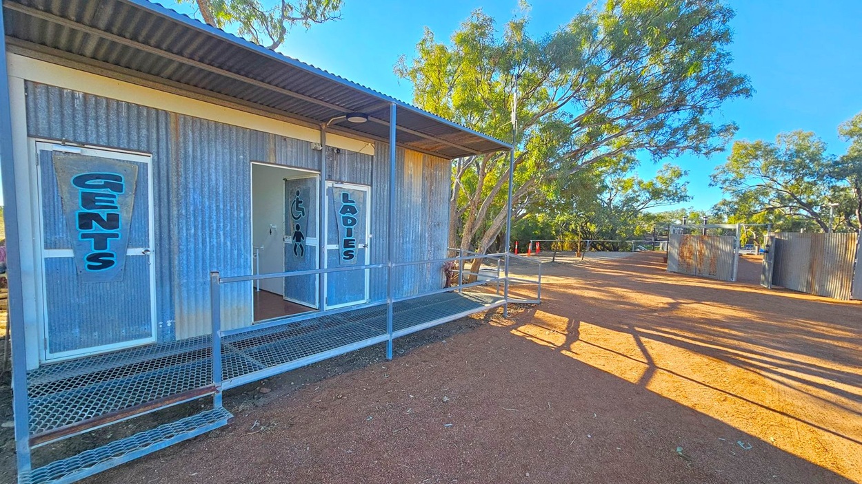 A corrugated metal building with two doors labelled "Gents" and "Ladies" on the left. The building has a metal ramp leading to the entrances. The ground is covered with reddish-brown dirt. In the background, there are trees and additional metal structures. The area is well-lit, suggesting daytime. Shadows from the trees and structures stretch across the ground.