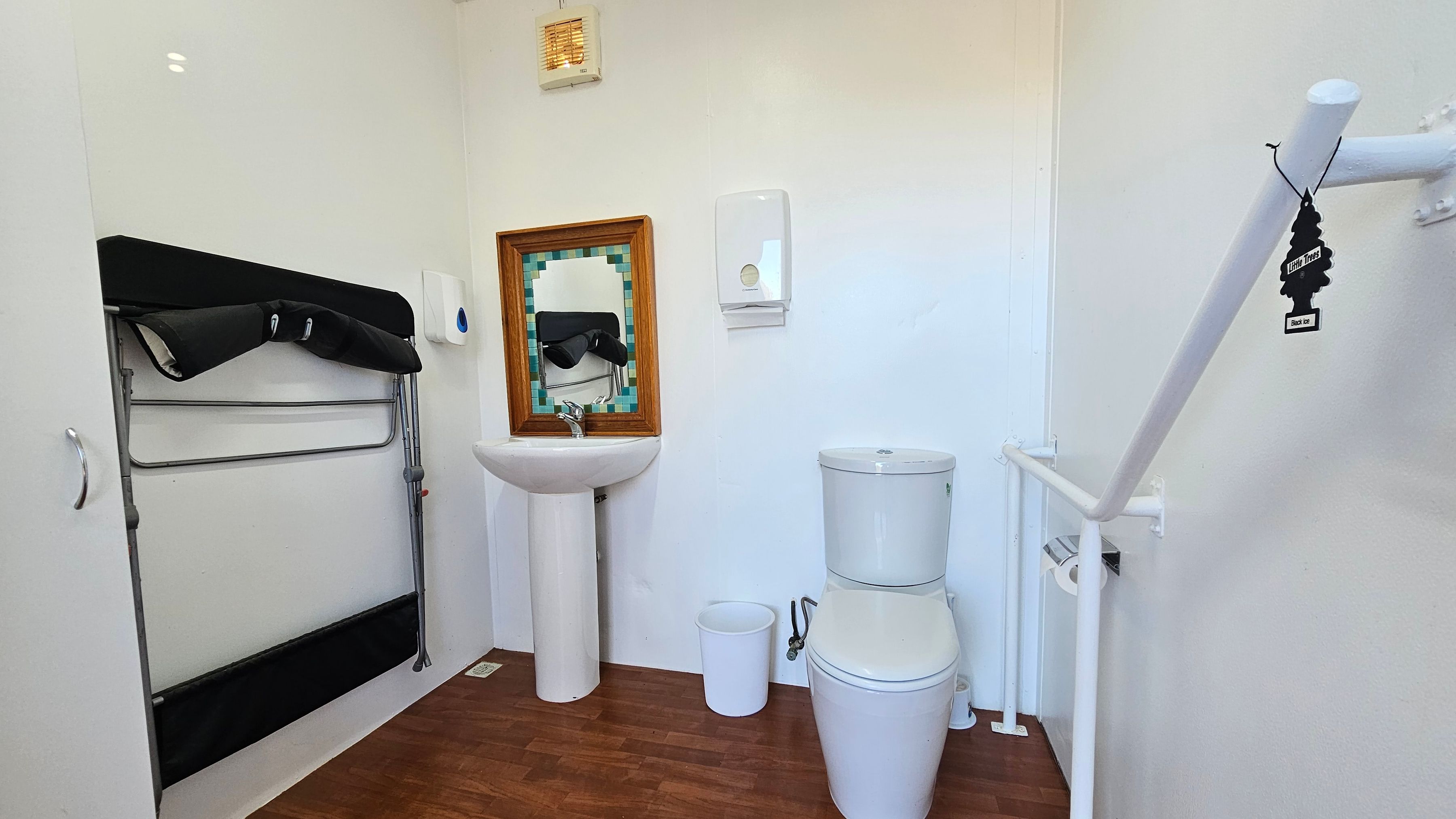 A bathroom with a wooden floor. On the left, a foldable black changing table is mounted on the wall. In the centre, a white pedestal sink with a wooden-framed mirror above it and a soap dispenser to the right. A white toilet is next to the sink, with a small white bin beside it. On the right, a white grab rail is attached to the wall, with a black tree-shaped air freshener hanging from it. The walls are light-coloured, and the ceiling has a small vent.