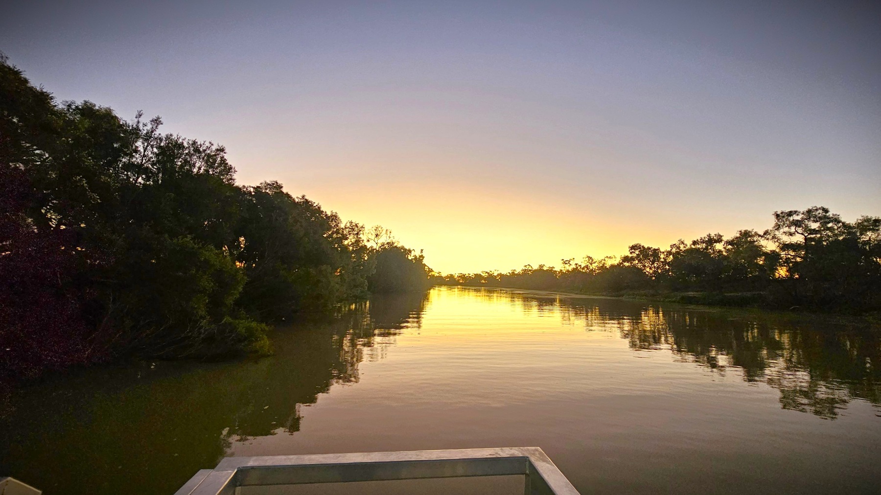 A calm river stretches into the distance, flanked by dense trees on both sides. The water reflects the soft light of the setting or rising sun, creating a serene atmosphere. In the foreground, a corner of a structure, possibly a dock or platform, is visible. The sky transitions from a pale yellow near the horizon to a deeper purple above, adding to the tranquil scene.