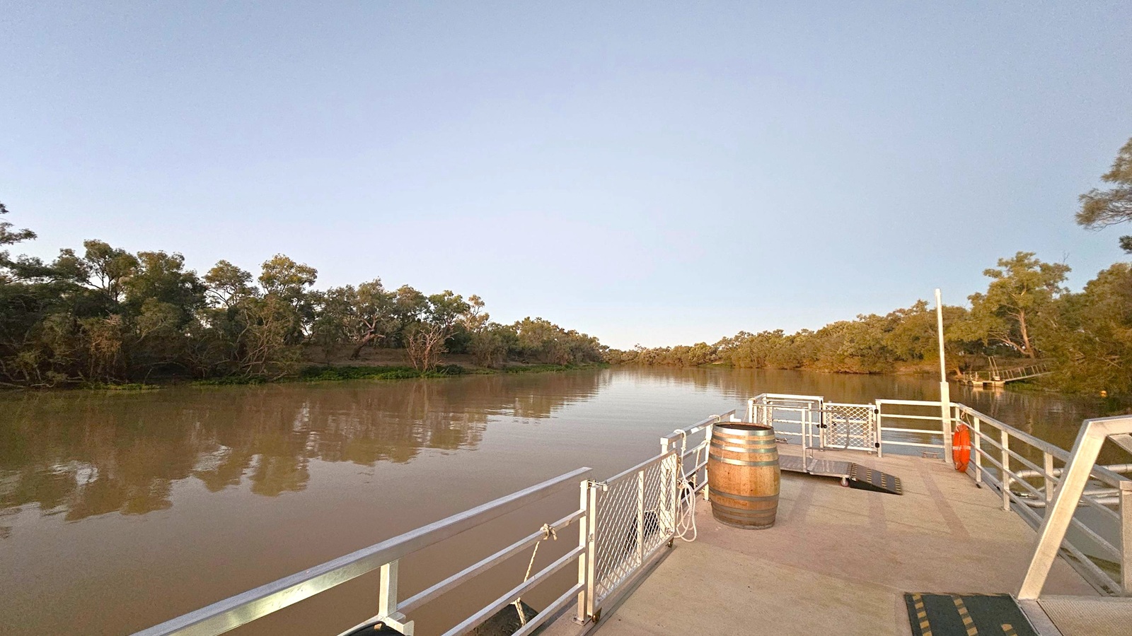 A wide river with calm, brownish water reflecting the surrounding trees. On the left, dense greenery lines the riverbank. On the right, a metal platform extends over the water, featuring a wooden barrel, a lifebuoy, and safety railings. The platform has a non-slip surface with yellow and black markings. Trees are visible in the background on both sides of the river.