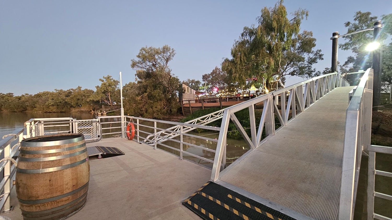 A wooden barrel is on the left side of a metal dock, with a life ring attached to the railing. A ramp with a metal frame leads up to the right, illuminated by a light post. In the background, there are trees and a glimpse of a building with lights, possibly part of a fair or event. The water is visible on the left, bordered by more trees. The dock surface is concrete, and the ramp has a textured surface.