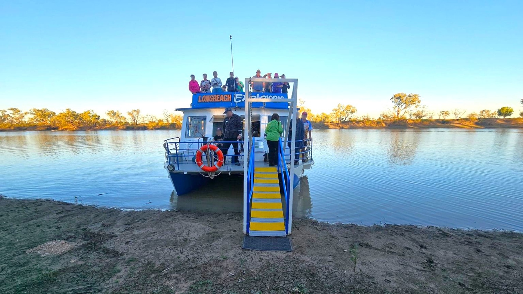 A boat named "Longreach" is docked on a calm body of water. The boat has a blue and white exterior with a group of people on the upper deck and a few on the lower deck. A yellow and blue gangway extends from the boat to the muddy shore. In the background, there are trees lining the opposite bank, reflecting in the water. The sky is clear, and the scene is well-lit, suggesting daytime.