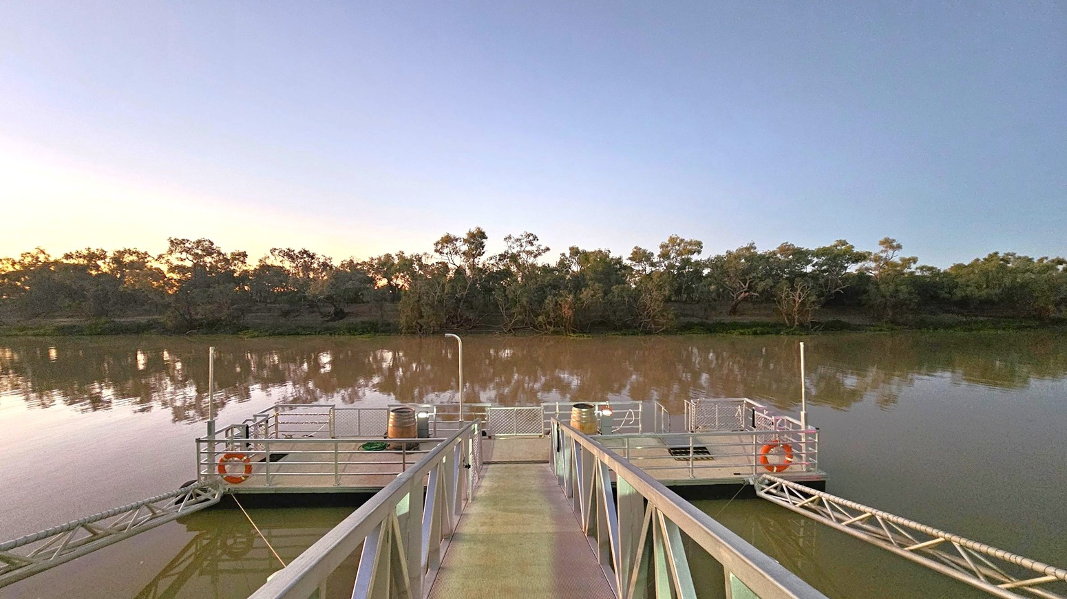 A metal walkway leads to a floating platform on a calm river. The platform has railings, life rings, and a few barrels. The river reflects the surrounding trees, which form a dense line along the opposite bank. The sky is a gradient of soft colours, suggesting early morning or late evening. The water is a muddy brown, and the trees are silhouetted against the sky.