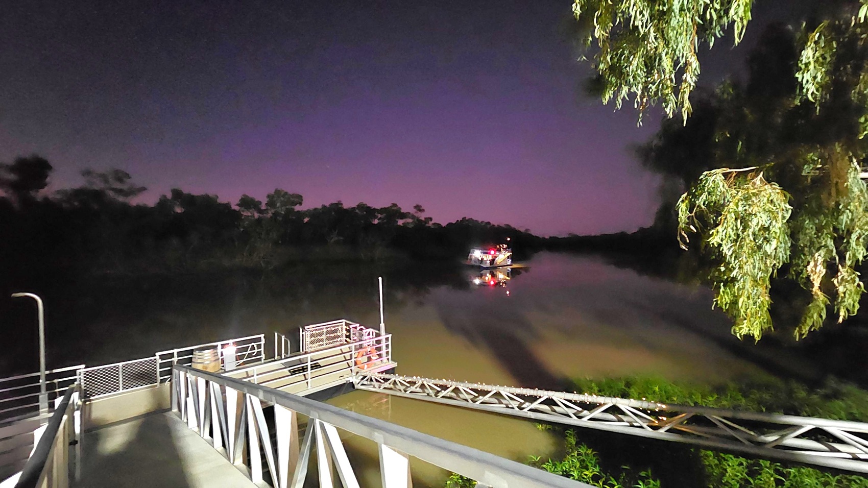 A metal walkway with railings leads down to a small dock on the left. The dock is illuminated with small lights. In the background, a boat with lights is visible on the water. On the right, there are overhanging tree branches. The water reflects the lights from the boat, and the surrounding area is dark, with silhouettes of trees against a dimly lit sky.