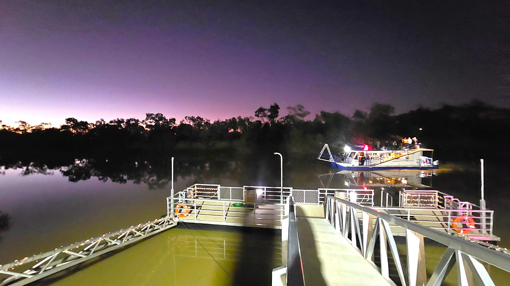 A river scene at dusk with a metal walkway in the foreground leading to a dock. The dock is equipped with safety railings and life rings. On the right, a boat with lights is moored, reflecting on the calm water. The background features silhouetted trees against a darkening sky with a gradient of purple and orange hues. The water is still, mirroring the trees and the boat.