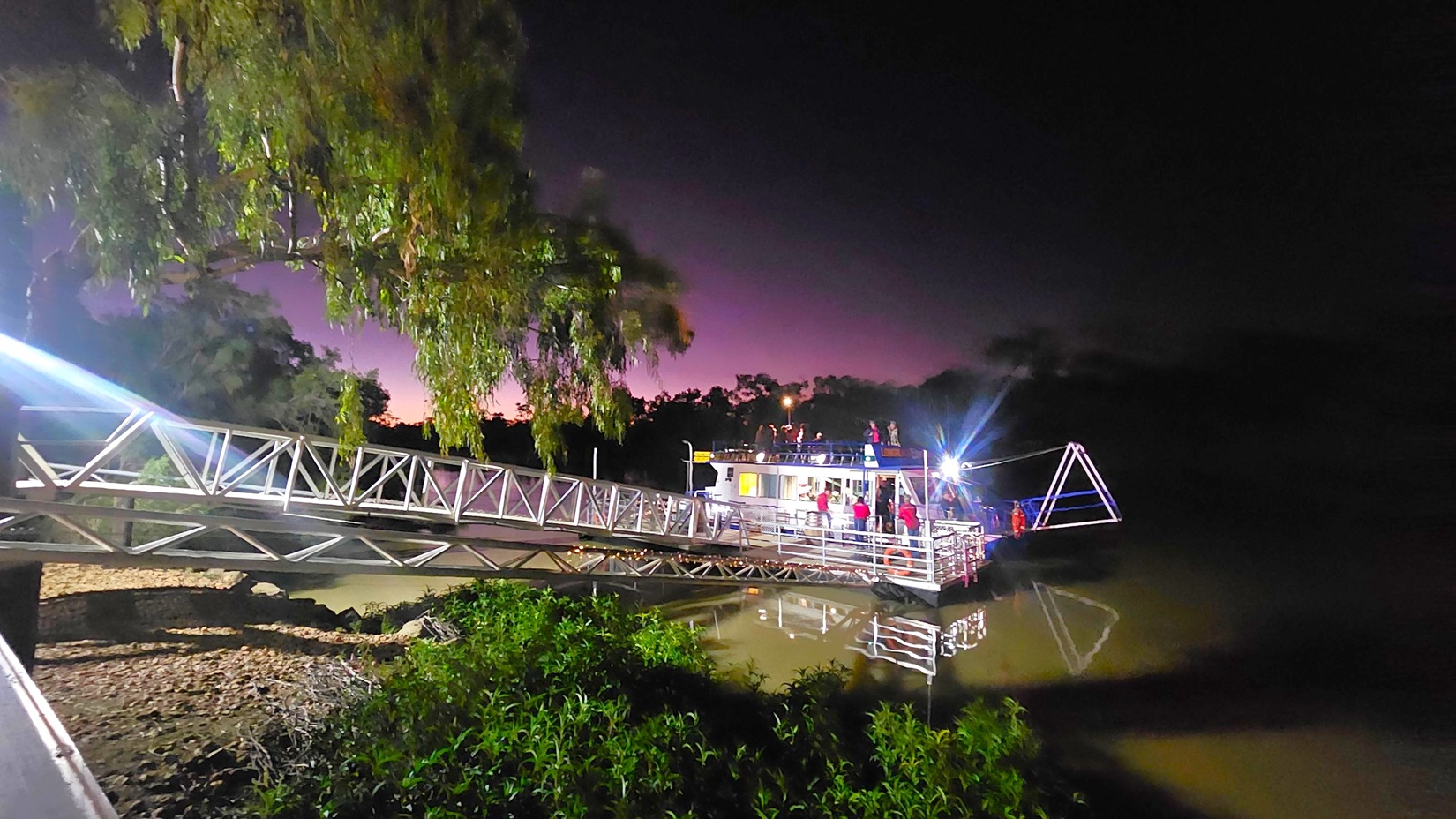 A metal walkway extends from the left, leading to a boat on the right. The boat is illuminated, with people visible on board. Trees with dense foliage are on the left, and the water below reflects the structure and lights. The sky in the background has a gradient from dark to a faint purple hue. Vegetation is visible in the foreground near the water's edge.