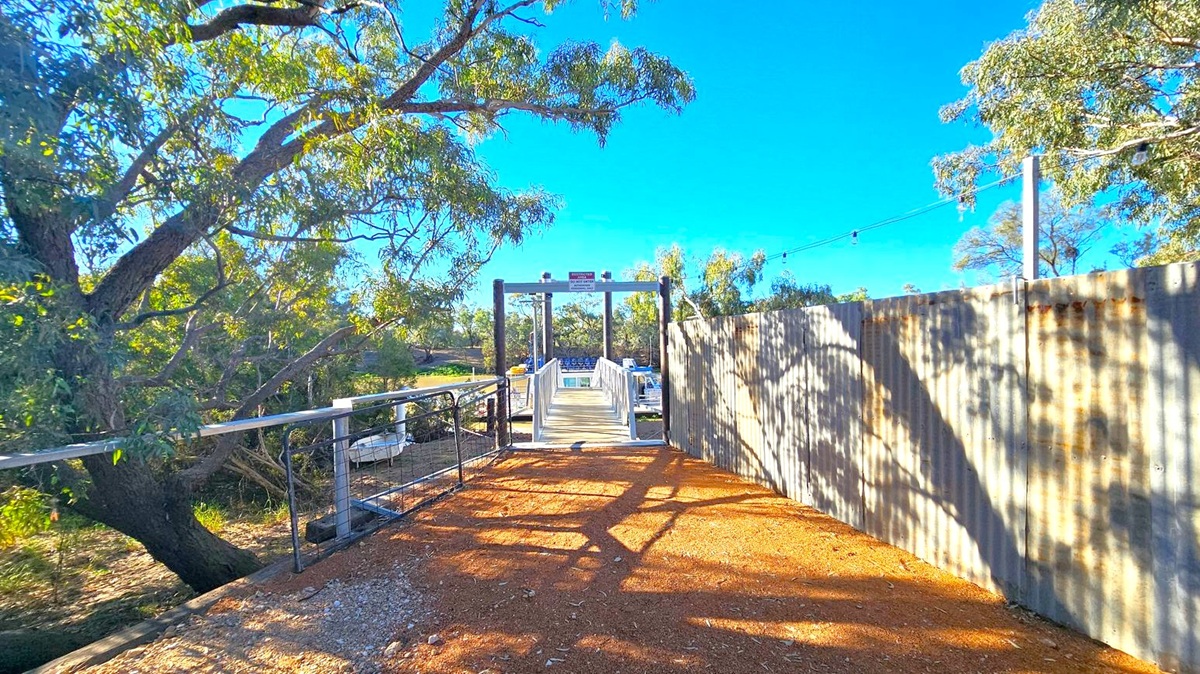 A pathway with a reddish-brown gravel surface leads to a small bridge in the centre. On the left, there is a large tree with overhanging branches casting shadows on the path. On the right, a corrugated metal fence runs alongside the path, also casting shadows. In the background, more trees are visible, and the bridge has a sign on top. The sky is clear and blue.
