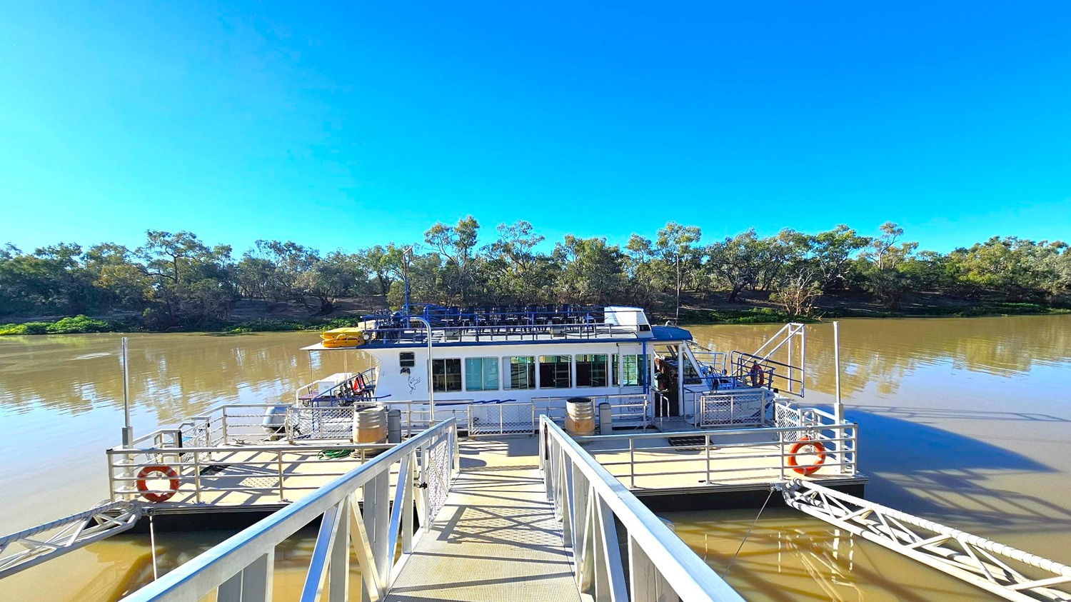 A boat is docked on a river, with a metal walkway leading to it. The boat has a white exterior with windows and a rooftop seating area. Yellow life rafts are visible on the deck. The river is calm, with trees lining the opposite bank. The sky is clear and blue.