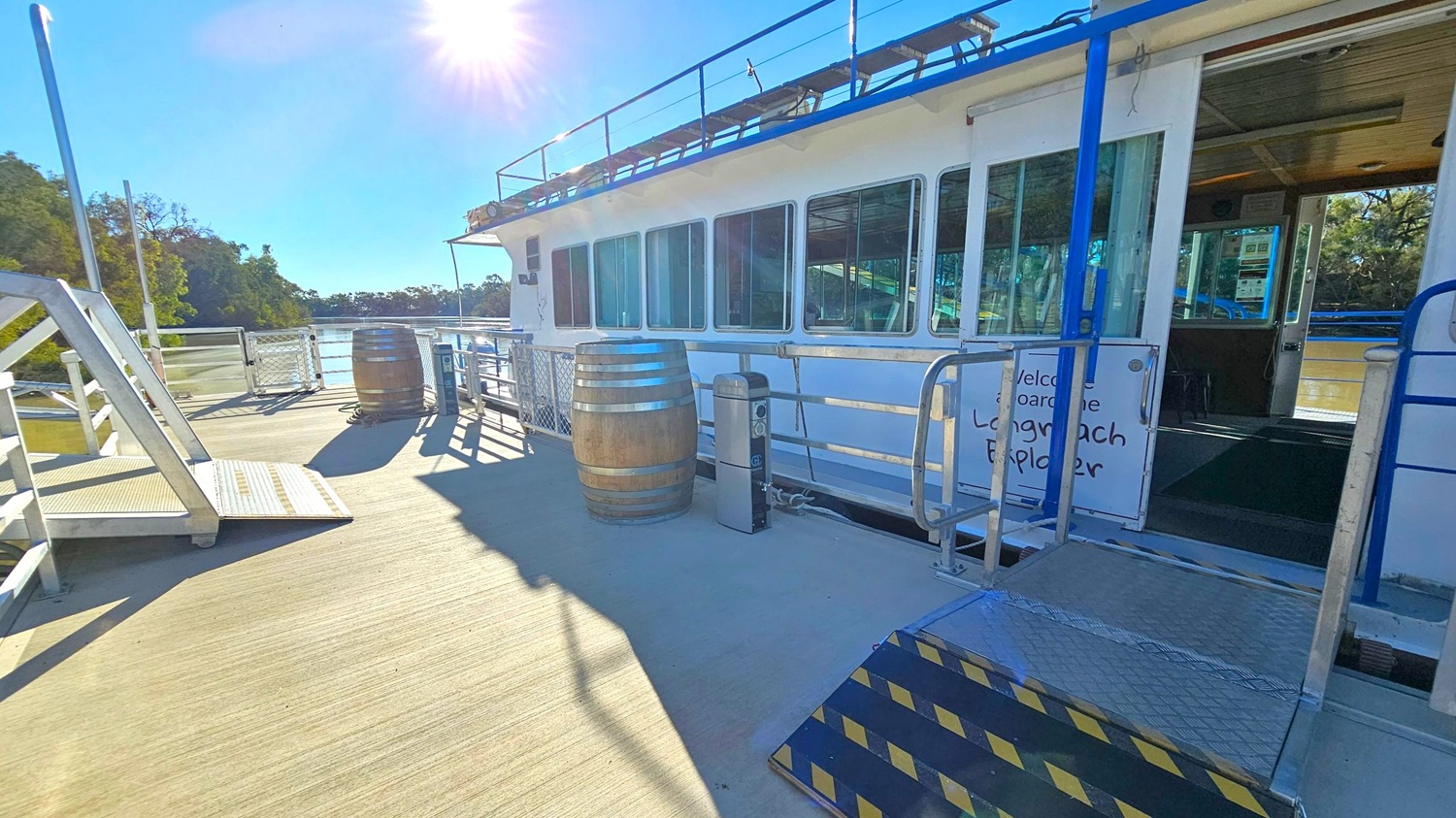 A boat docked at a pier with a ramp leading to it. The boat has large windows and a sign that reads "Welcome aboard the Longreach Explorer." Two wooden barrels are placed on the pier, along with a small metal post. The sun is shining brightly in the sky, casting shadows on the wooden deck. Trees and water are visible in the background.