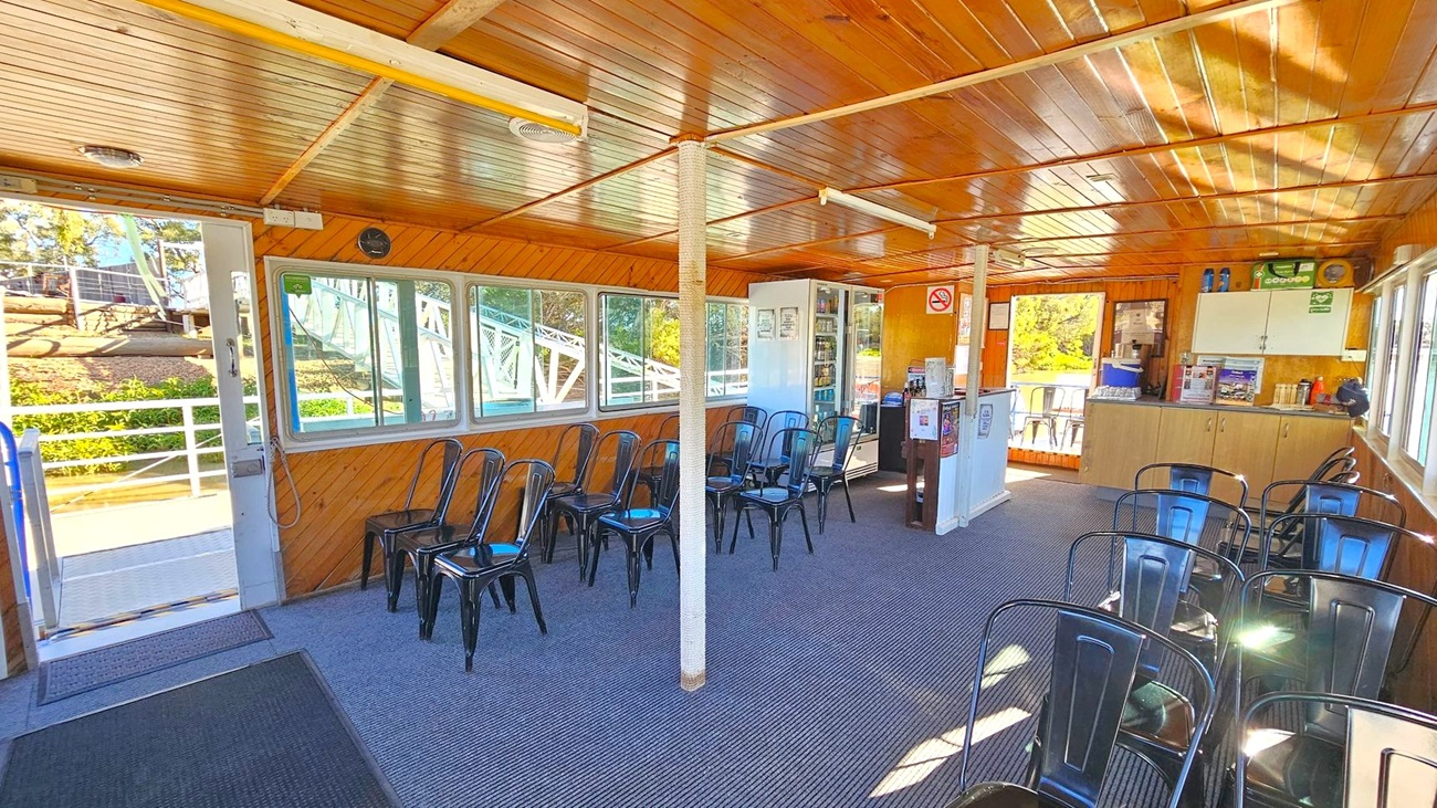 Interior of a wooden cabin with a slatted ceiling and carpeted floor. On the left, a door opens to an outdoor area with a view of trees and a railing. Several black metal chairs are arranged along the walls and in the centre. On the right, a counter with cabinets above and a small fridge below holds various items. Windows line the walls, allowing natural light to illuminate the space. A white column stands in the middle of the room.