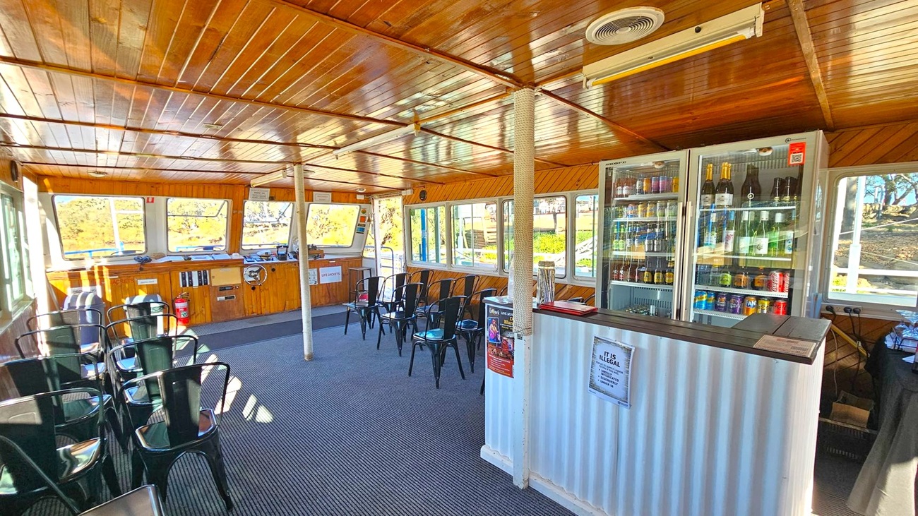 Interior of a boat with a wooden ceiling and walls. On the right, a bar area with a corrugated metal counter and a glass-fronted fridge stocked with drinks. In the middle, a row of black metal chairs on a carpeted floor. On the left, large windows provide views of the outside, with more chairs arranged along the wall. A fire extinguisher and life jacket sign are visible near the helm. The space is well-lit with natural light streaming through the windows.