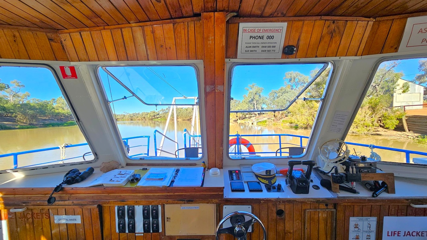The image shows the interior of a boat's control cabin. The foreground features a wooden dashboard with various controls, switches, and navigation equipment. Papers, a pair of binoculars, and a small fan are also visible. Through the large windows, a river with trees lining the banks can be seen. A life ring is attached to the boat's railing outside. The ceiling is wooden, and emergency contact information is displayed above the windows.