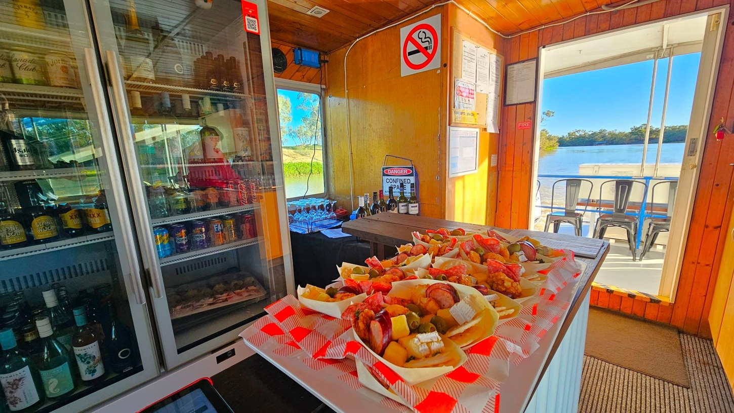 Inside a wooden cabin, a counter on the left holds a glass-fronted fridge stocked with various drinks. In the centre, a table displays trays of appetisers with meats and cheeses. The right side features an open door leading to a view of a river and outdoor seating with metal chairs. Walls have signs and notices, including a no-smoking sign. The floor is carpeted, and the ceiling is wooden.